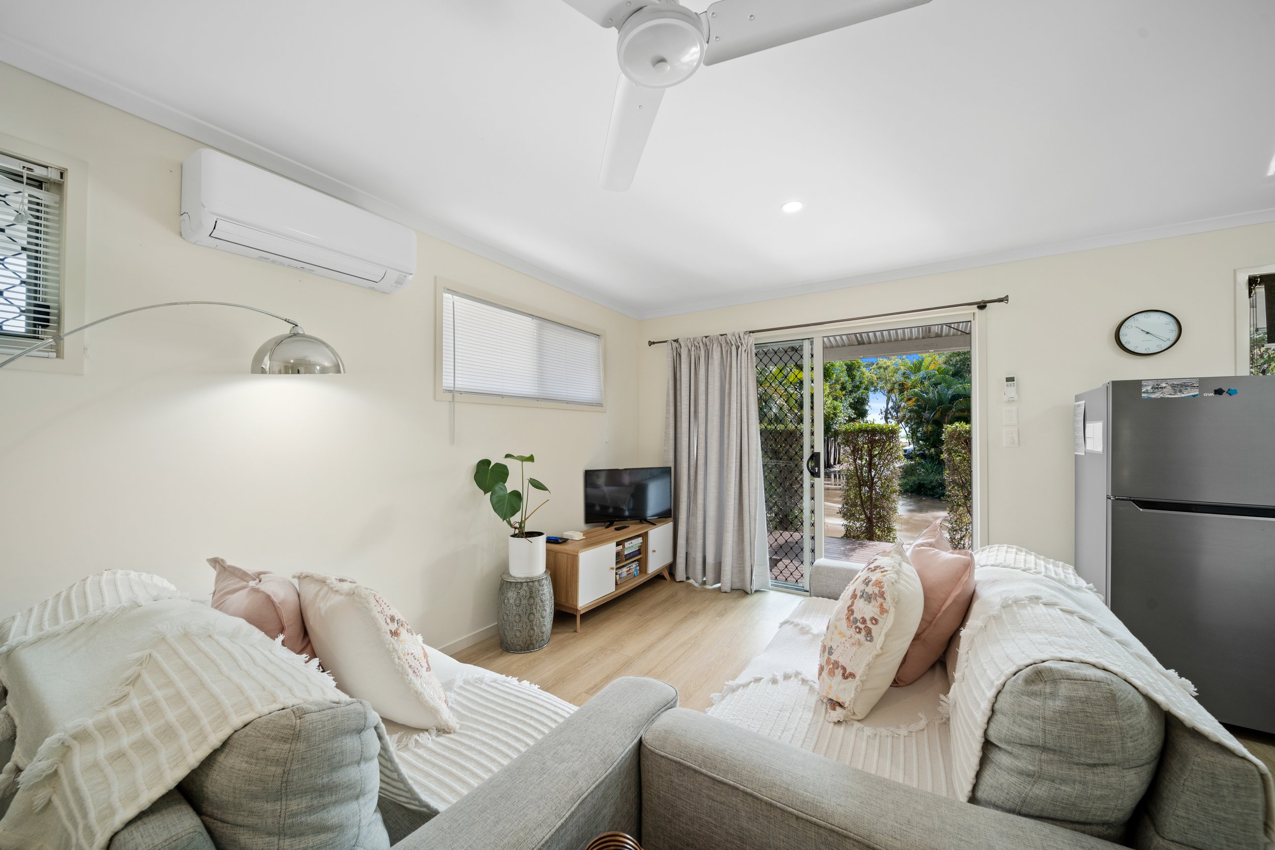 Living room with white walls and ceiling, ceiling fan, air conditioning unit, window with blinds, sliding glass door with curtains leading to outdoor patio, small TV on wooden stand, potted plant, clock on wall, closed refrigerator, and cream-colored