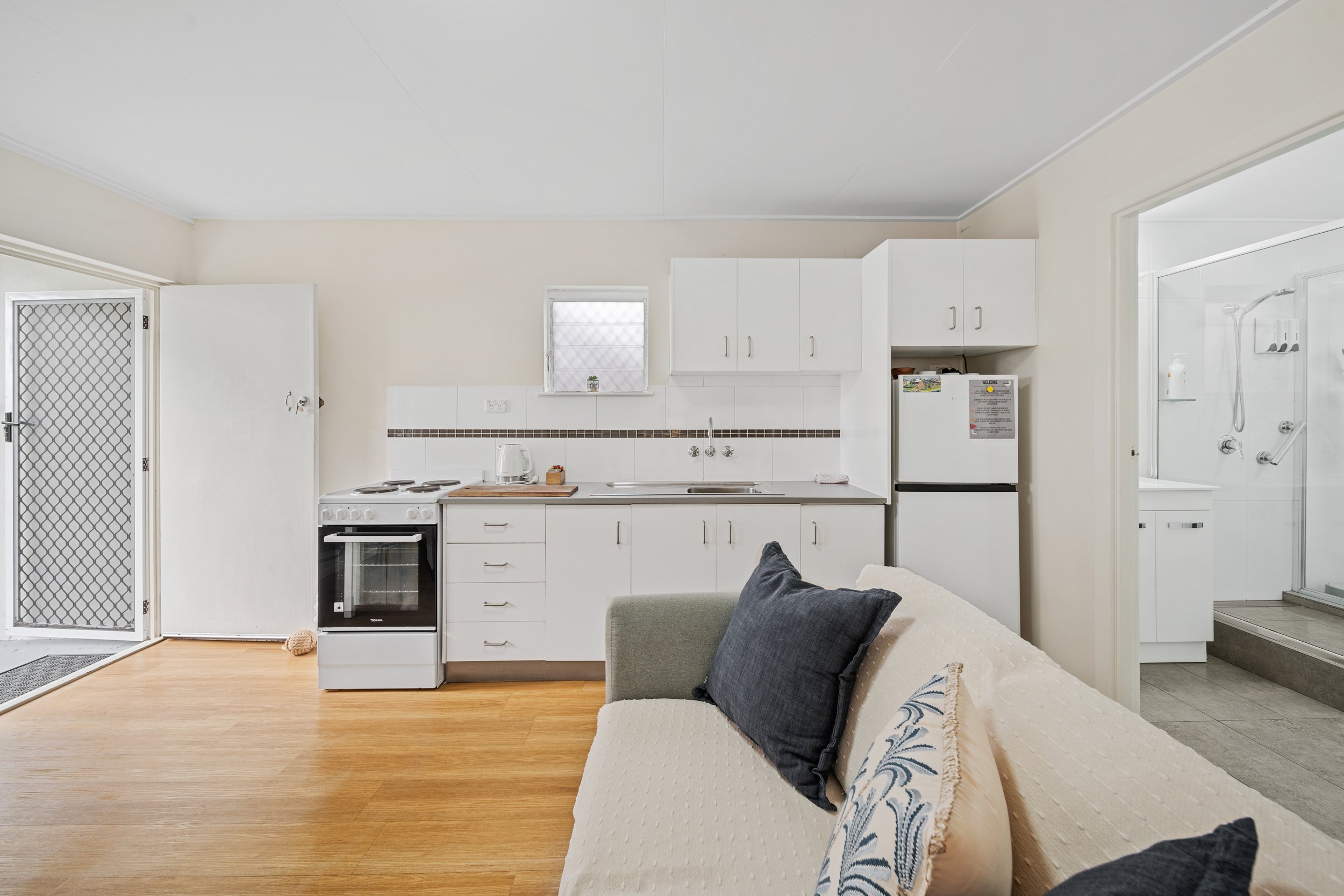 Interior view of a small kitchen with white cabinets, a stove, a microwave, and a refrigerator. There is a window above the sink and a door with a screen on the left. A beige couch with black and cream pillows is partially visible in the foreground.