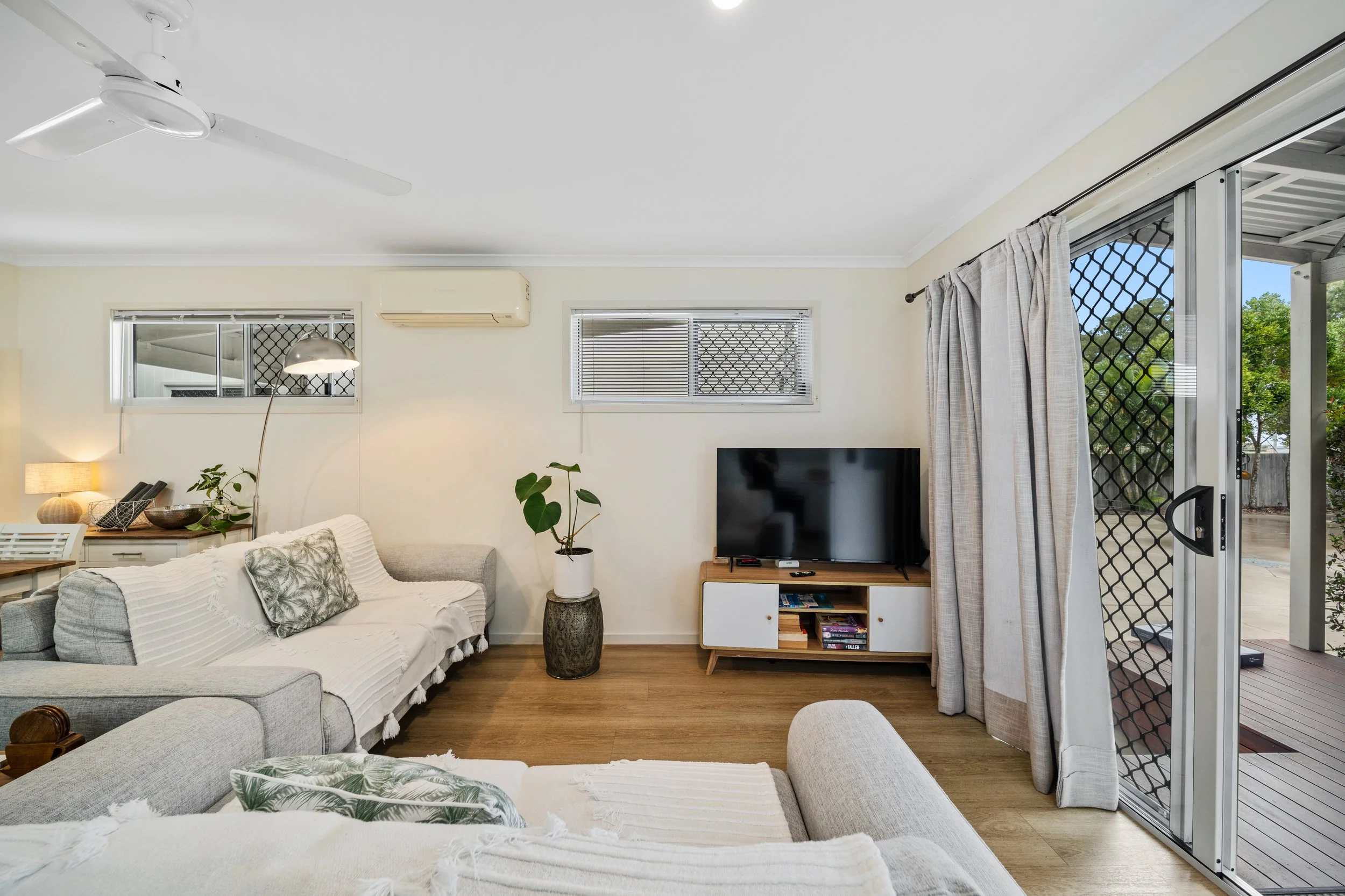 Living room with beige sofas, a wooden coffee table, potted plants, a wall-mounted TV, large sliding glass door with curtains, and windows with blinds, opening to an outdoor patio.