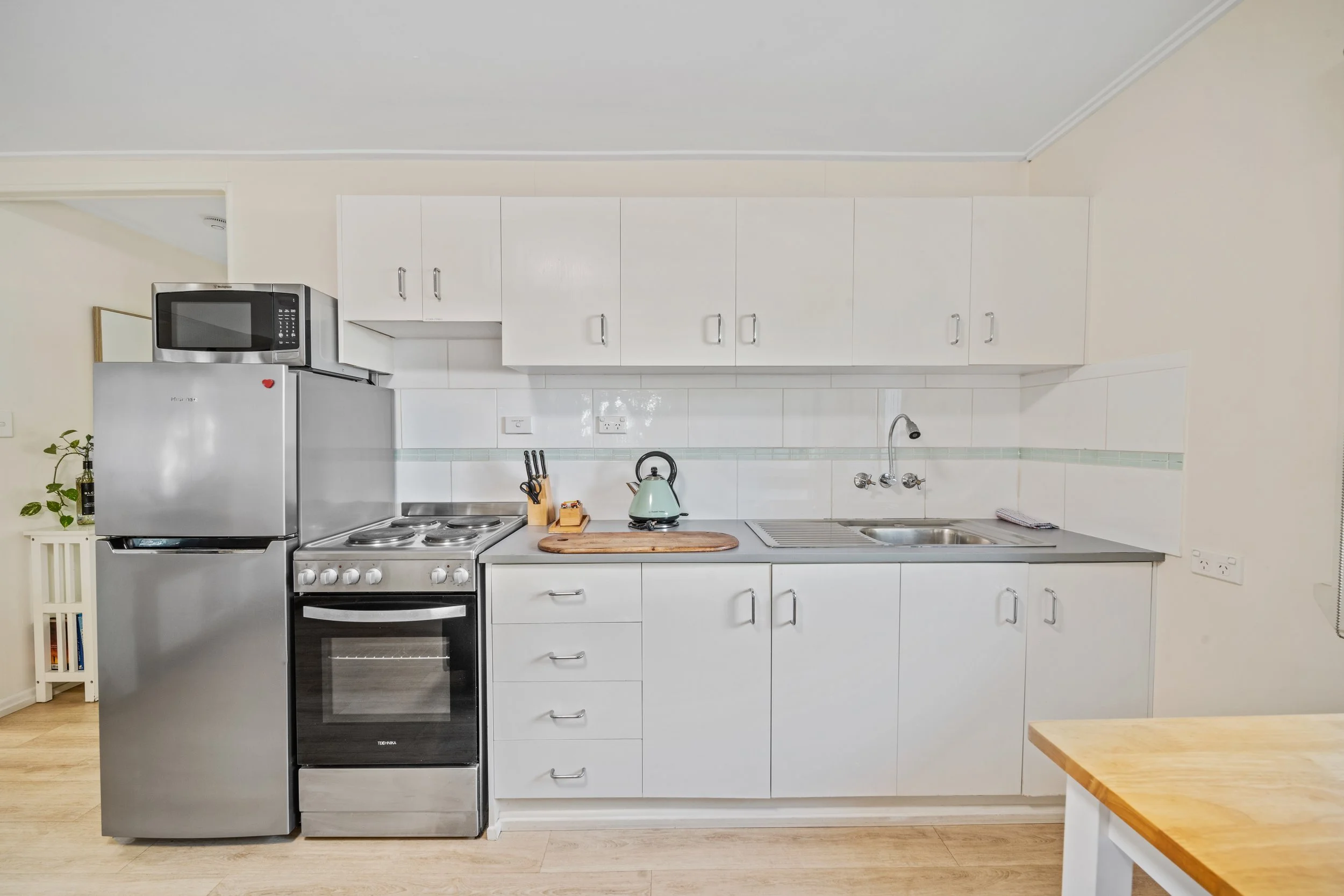 A kitchen with white cabinets, a stove, a microwave, a refrigerator, a kettle, a cutting board, and a sink.