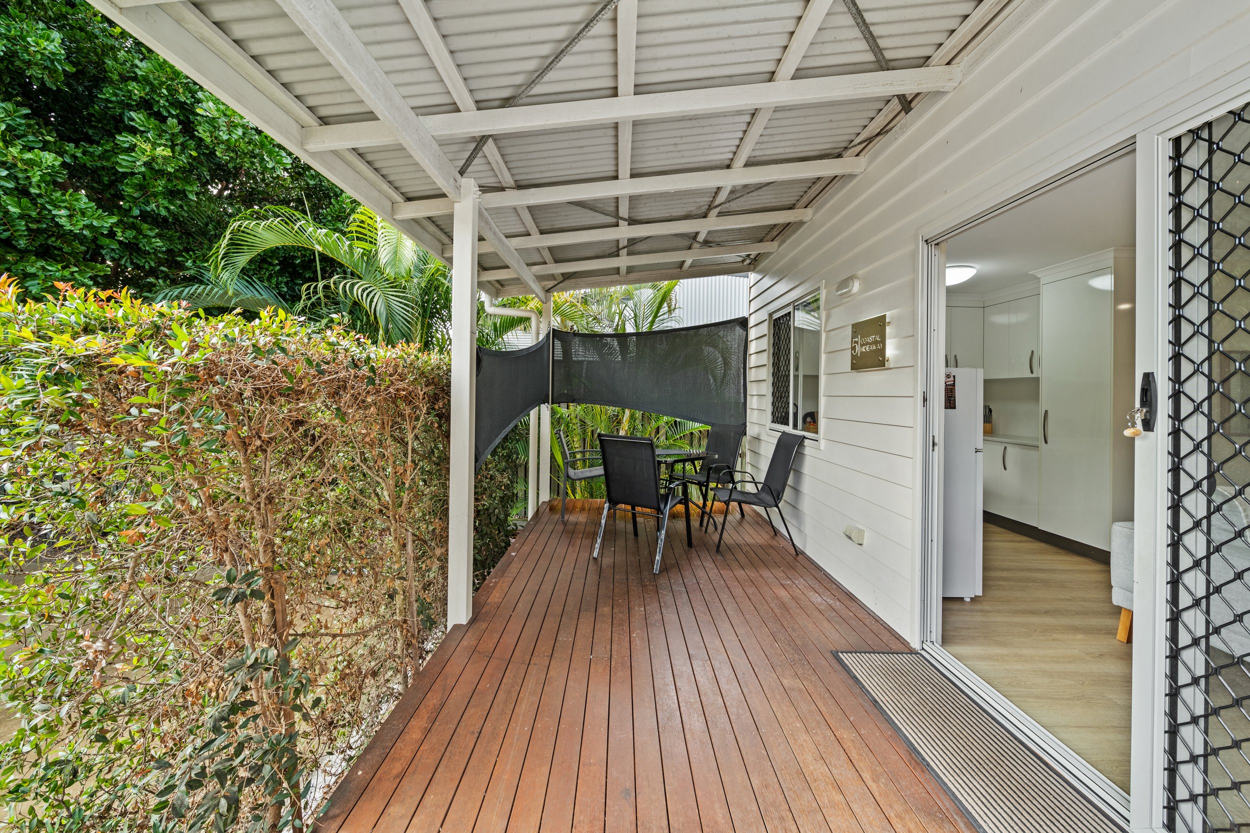 A covered outdoor patio with wooden decking, black chairs, and surrounding greenery, attached to a house with a sliding glass door and white walls.