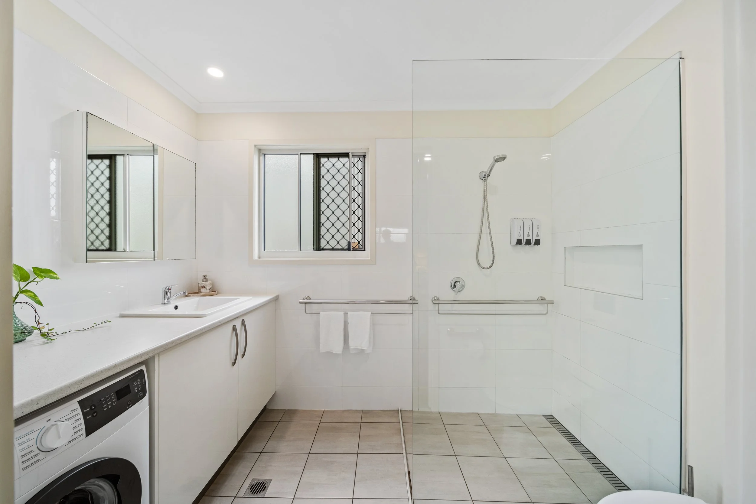 A modern bathroom with white tiled walls, a window with security bars, a walk-in shower area with a handheld showerhead, soap dispensers, a towel rail with a towel, a sink with a faucet, a mirror, a potted plant on the countertop, and a washing machi