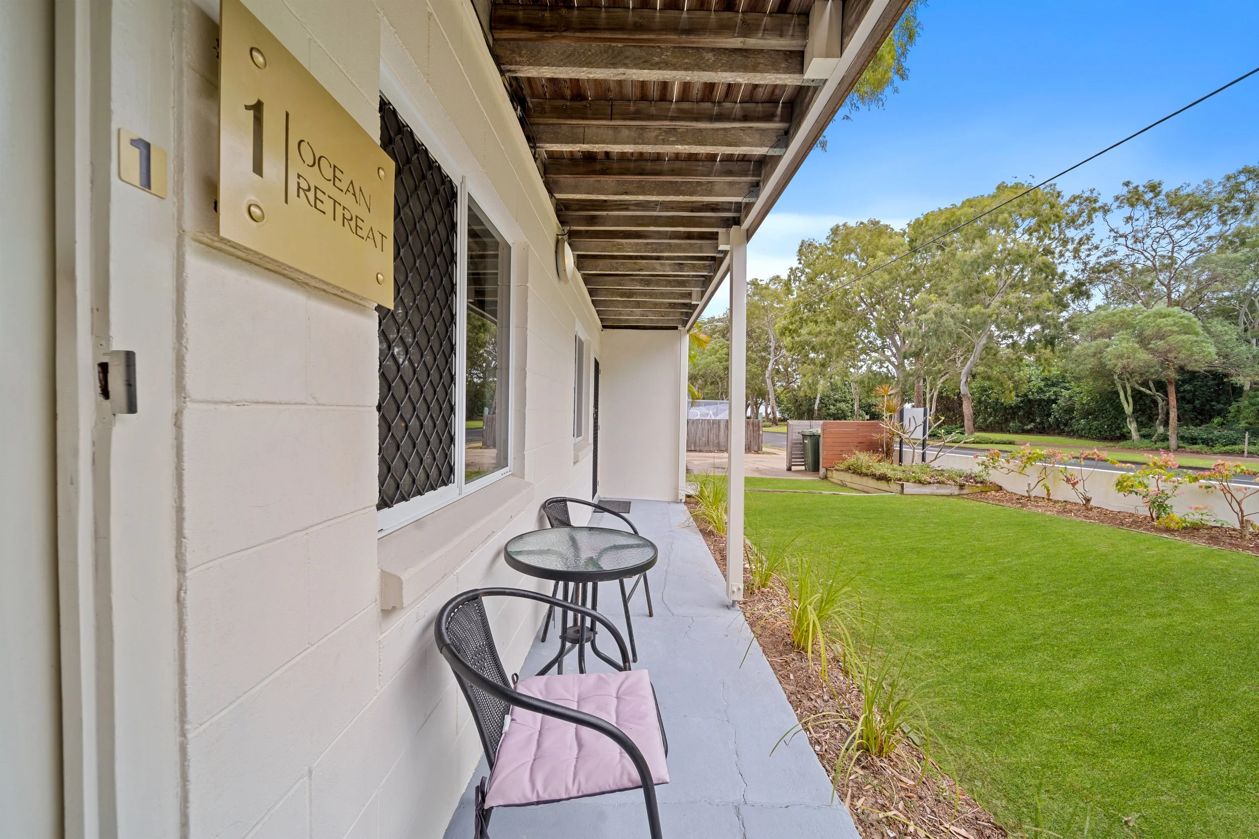 A small porch with black chairs and a pink cushion, attached to a white building with a sign that reads 'Ocean Retreat'. The porch has a concrete floor and is partially covered by a wooden ceiling. A garden with green grass and trees is visible in th
