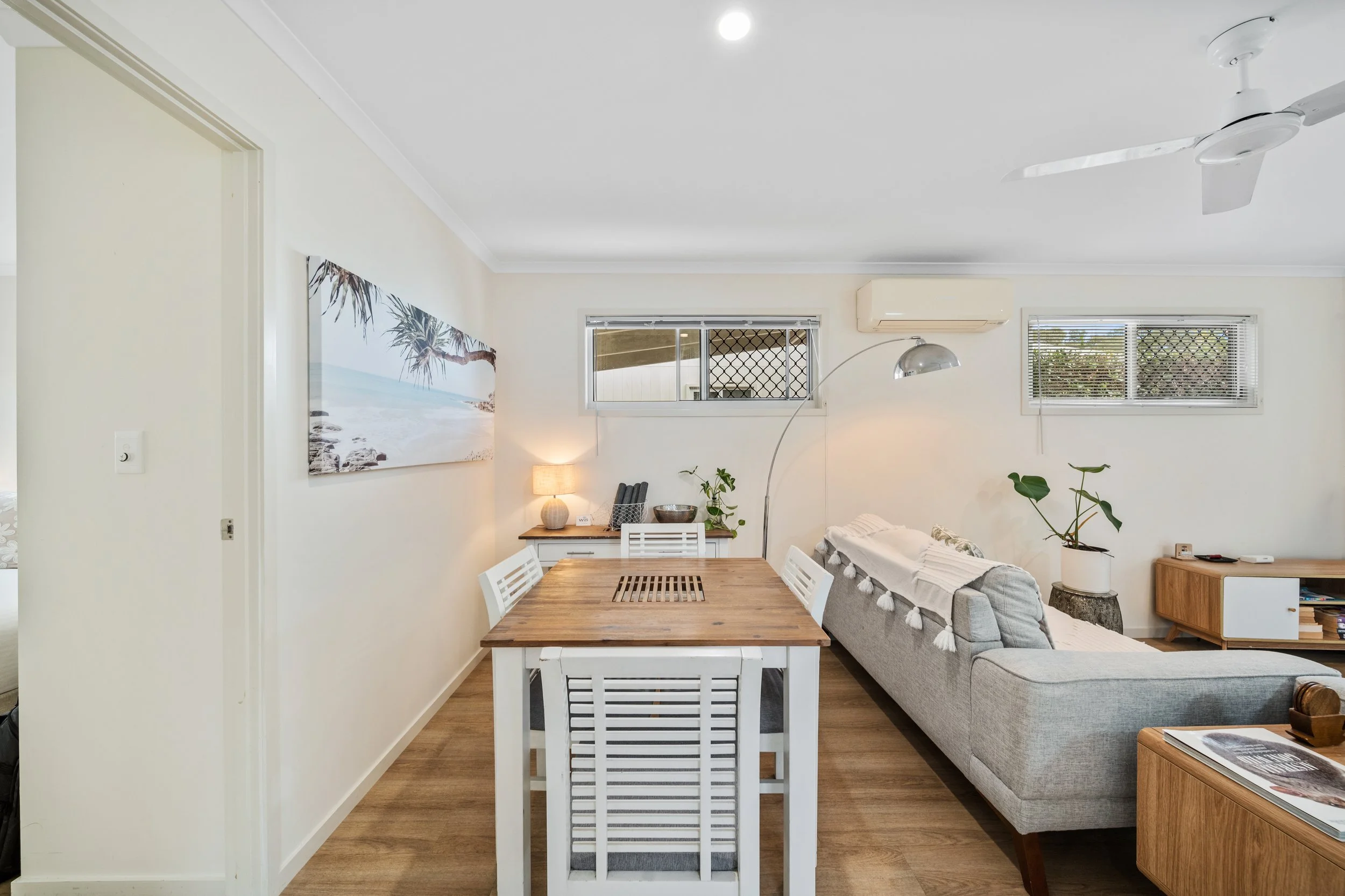 Interior of a living room and dining area with a wood dining table, white chairs, a gray sofa with decorative pillows, wall art of a beach scene, a lamp, indoor plants, and windows with blinds.