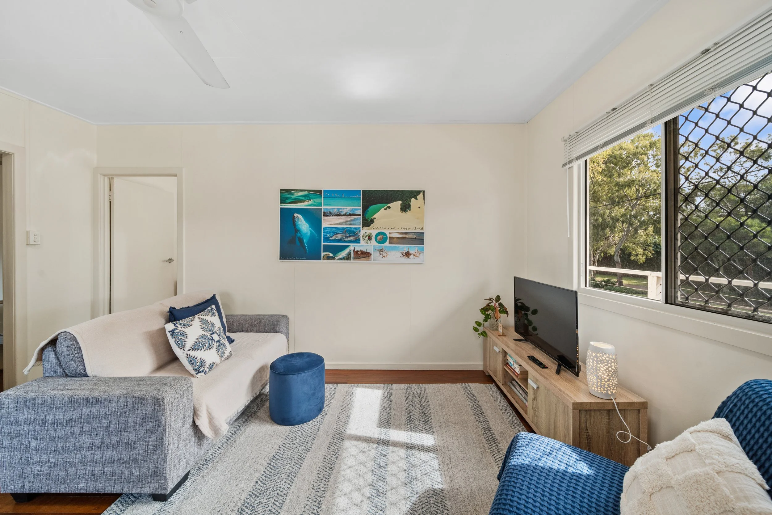 Living room with a gray sofa, blue and white throw pillows, a small round blue ottoman, a wooden TV stand with a television, a potted plant, and a window with white blinds showing trees outside. There is a collage of beach photos on the wall.