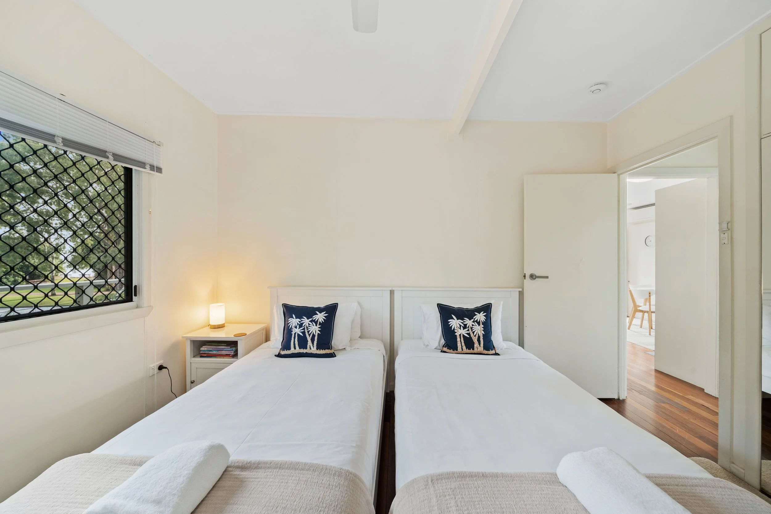A bedroom with two twin beds with white bedding and decorative pillows featuring palm trees. There is a nightstand with a lamp and books between the beds. A window with blinds and a view of greenery is on the left, and an open door leads to another r
