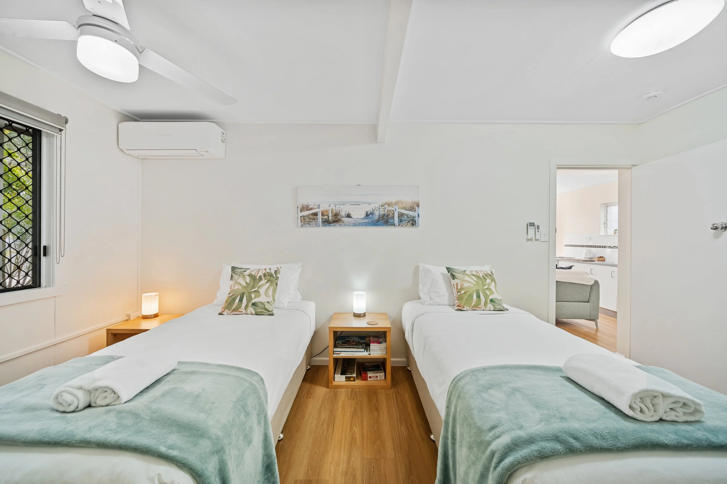 A bedroom with two single beds dressed in white and green bedding, each with a rolled towel, and small nightstands with lamps between them. There is a window with a view outside and a wall art depicting a beach scene. The room has wood floors, and in
