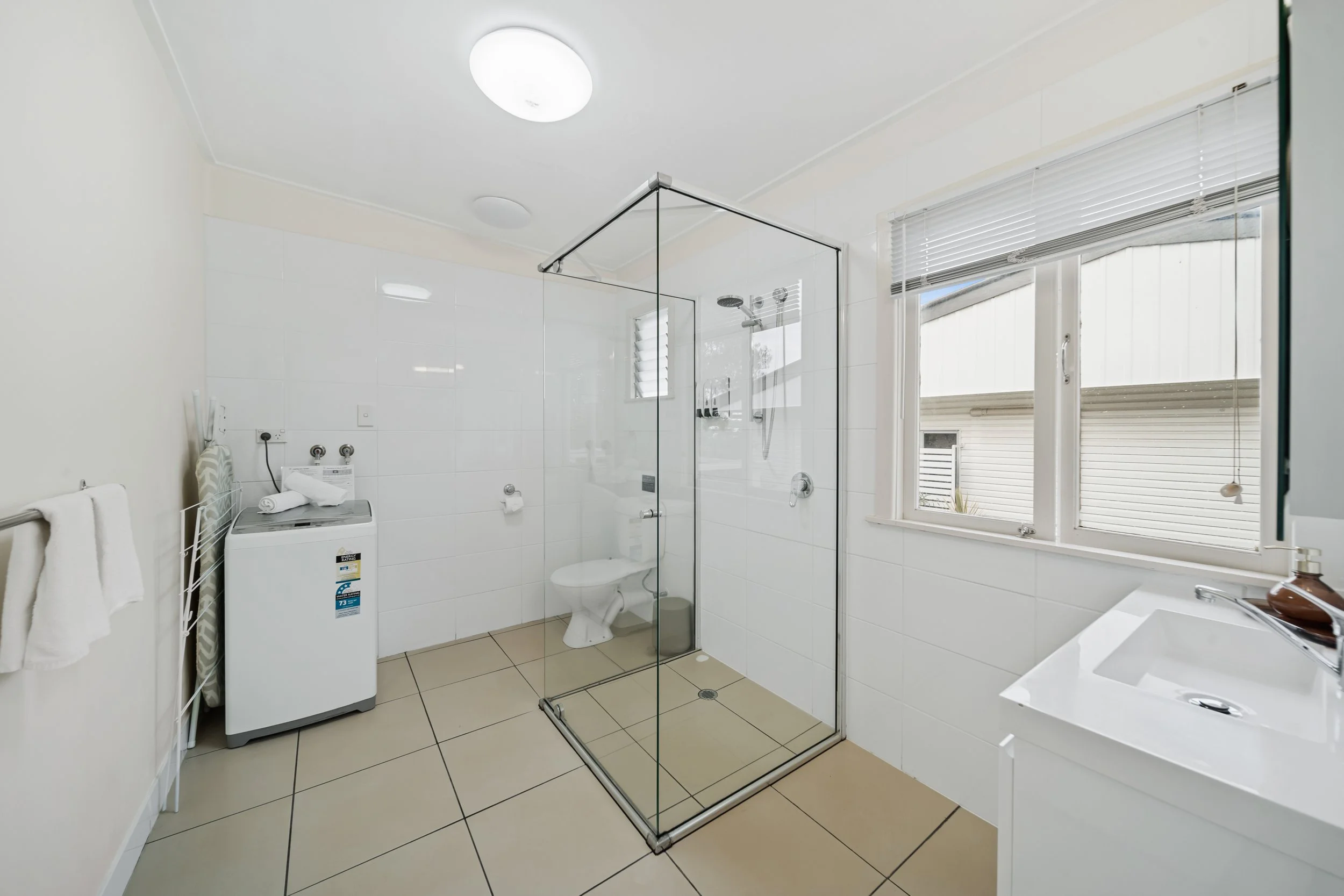 Bathroom with glass shower enclosure, white walls, window with blinds, washing machine, towel rack, and bathroom vanity.