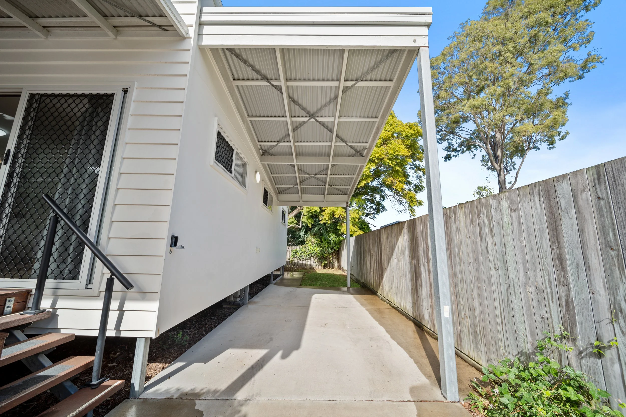 Backyard with a white house, a covered patio, a wooden fence, trees, and a concrete pathway.