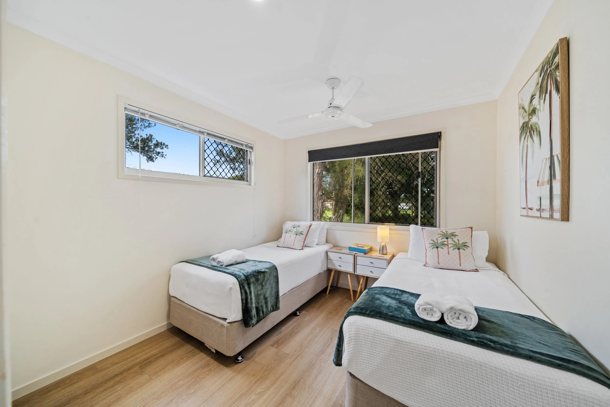 A bright bedroom with two single beds, each with white linens and decorative pillows with palm tree designs, a nightstand with a lamp and books, large windows with black security screens, a ceiling fan, and light-colored walls and wood flooring.
