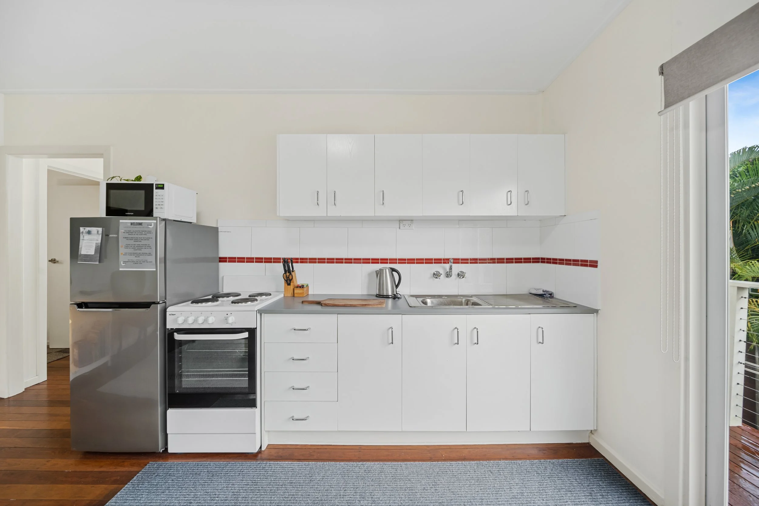 Kitchen with white cabinets, a stove, a microwave on top of a refrigerator, a kettle, a knife block, and a sink with a window showing outdoor greenery.