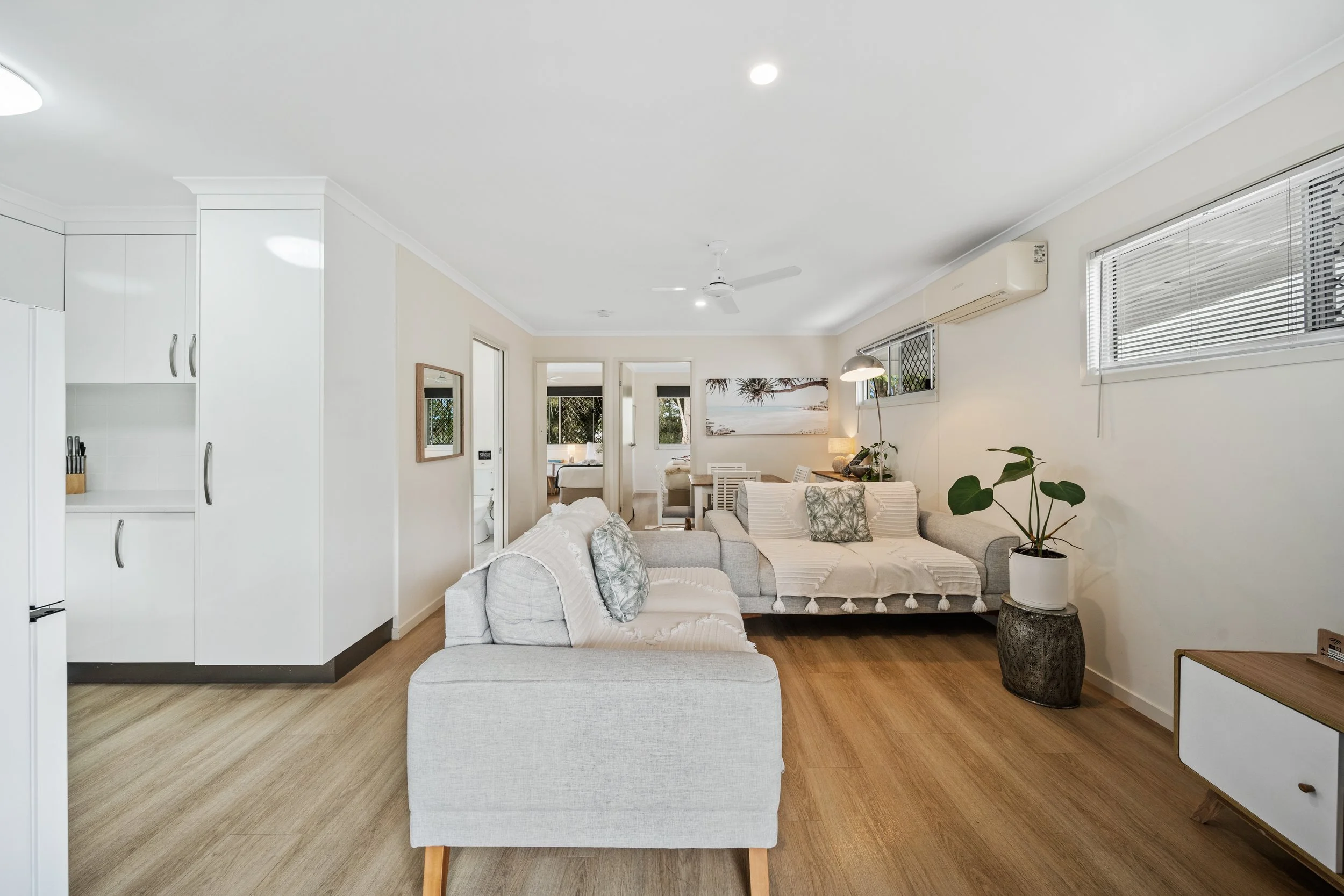 Living room with white walls, a gray sectional sofa with patterned pillows, a wooden table, a large potted plant, windows with blinds, an air conditioner, and a ceiling fan.