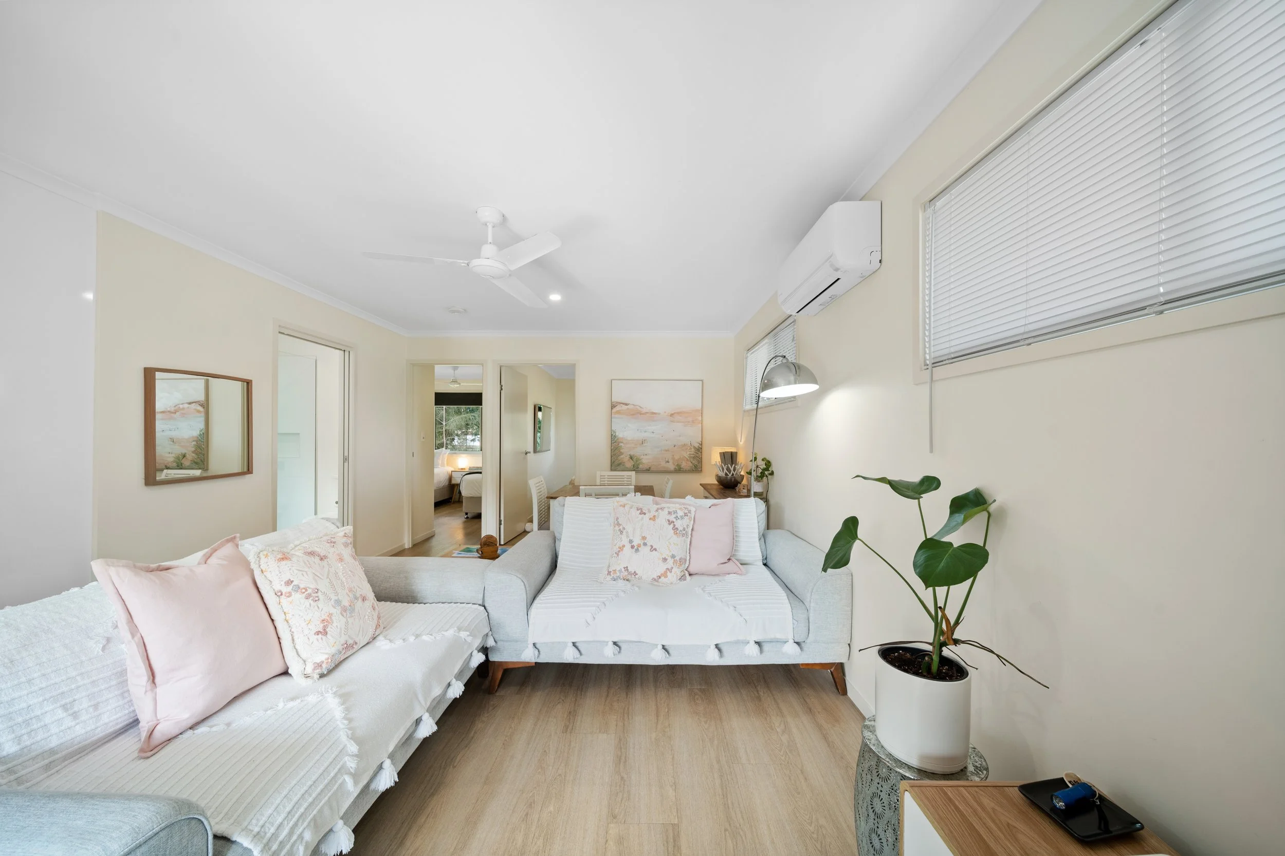 Living room with white sofa, pink pillows, potted plant, wall art, window blinds, and ceiling fan.