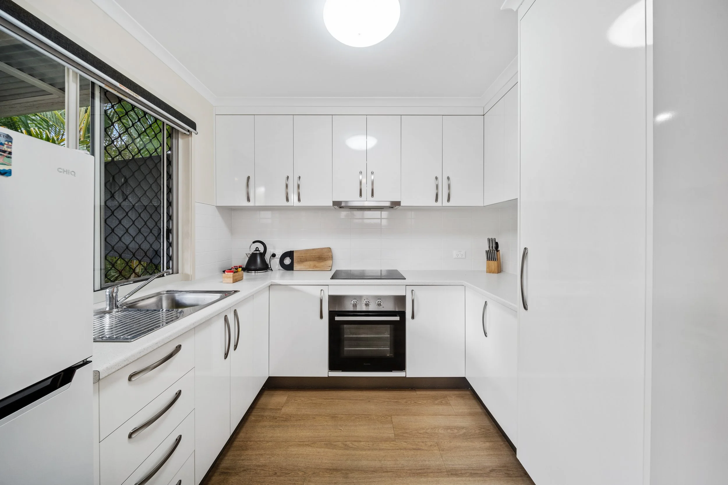 Clean, white kitchen with upper cabinets, a black stove, and wooden flooring. A window with a screen shows greenery outside. Kitchen utensils and appliances are neatly arranged.