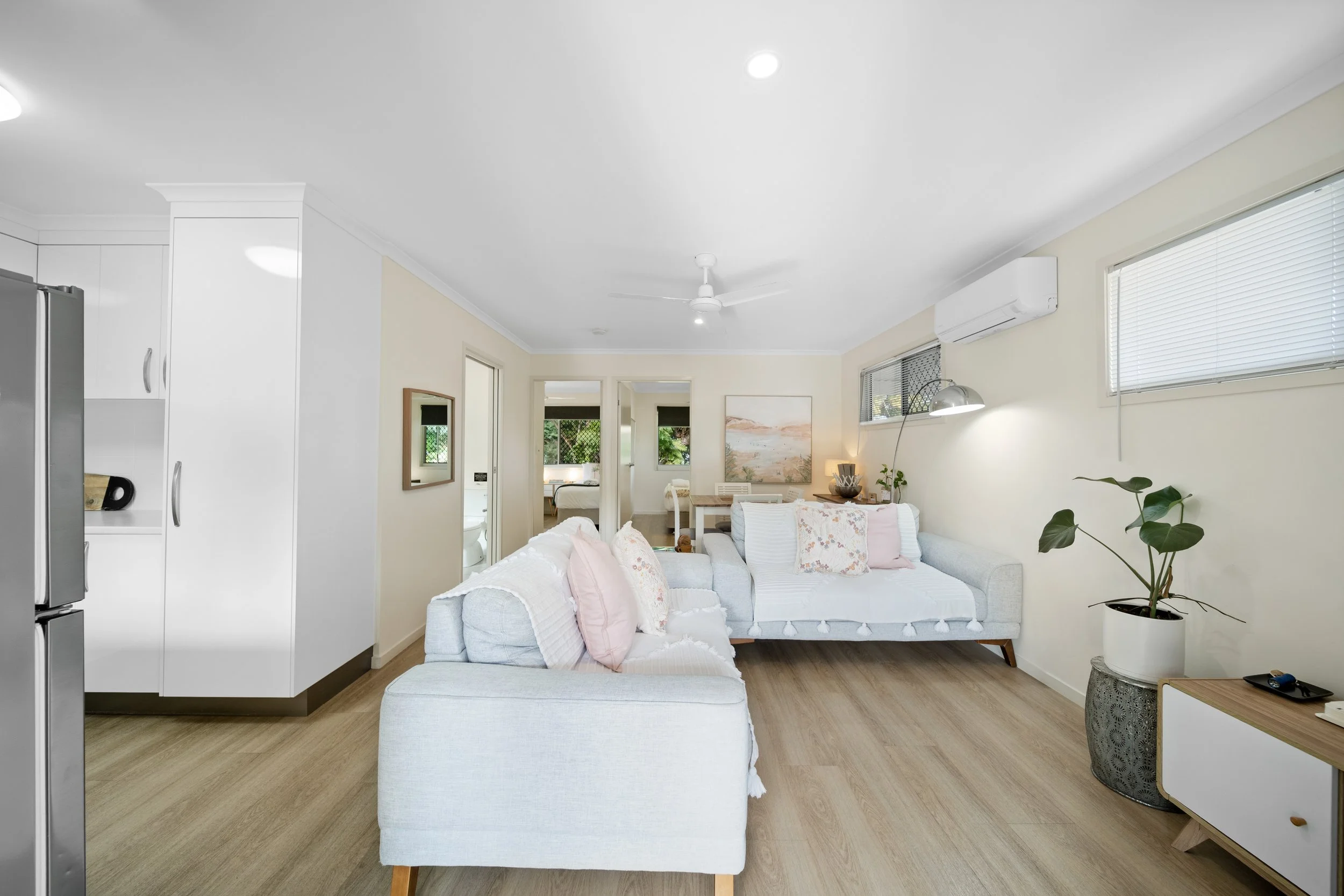 Bright living room with white sofa, pastel pillows, wooden flooring, and large windows with blinds. Open space with a small side table and potted plant.