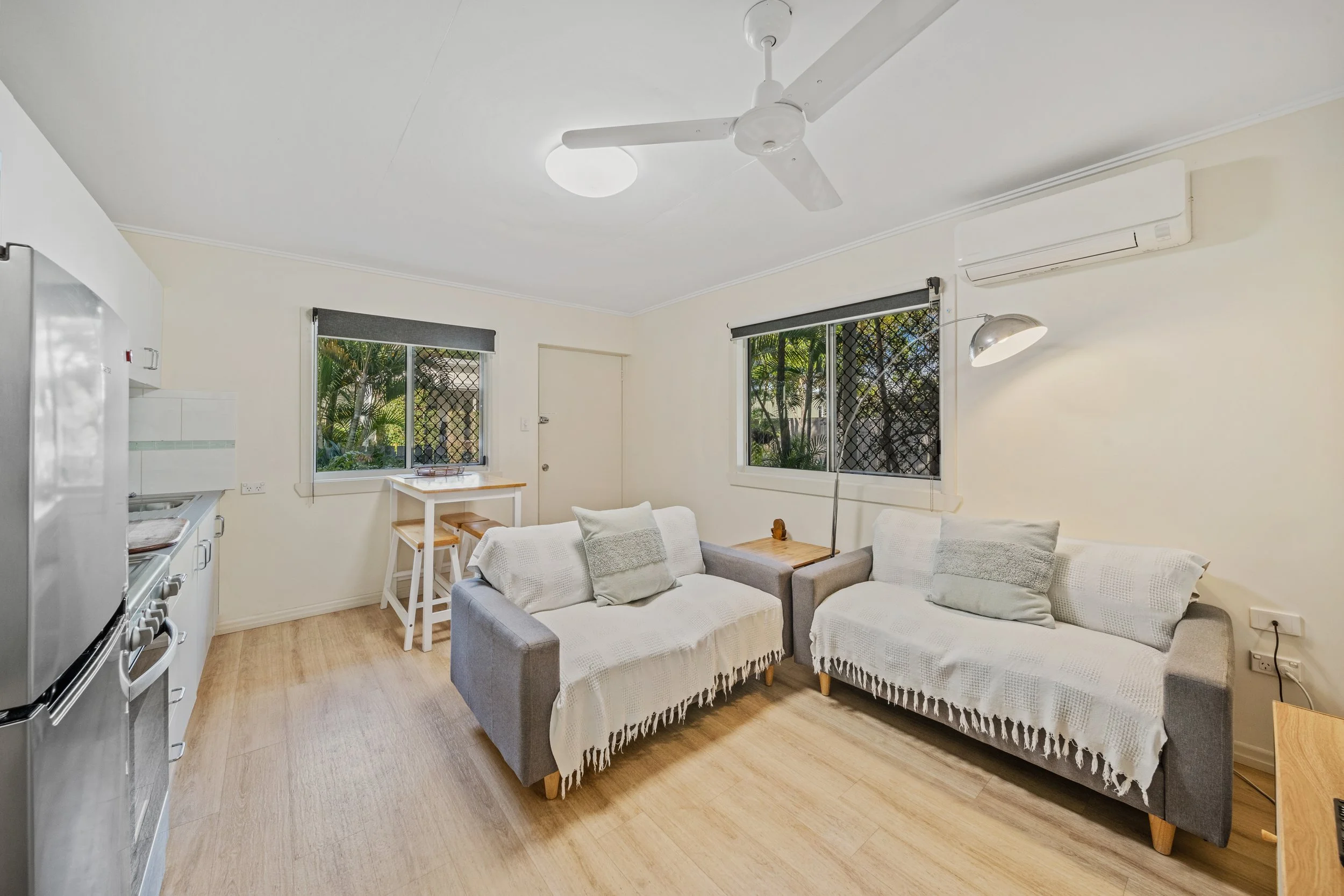 Living room with two couches, a small wooden dining table, windows with black mesh screens, ceiling fan, air conditioner, floor lamp, and wooden flooring.