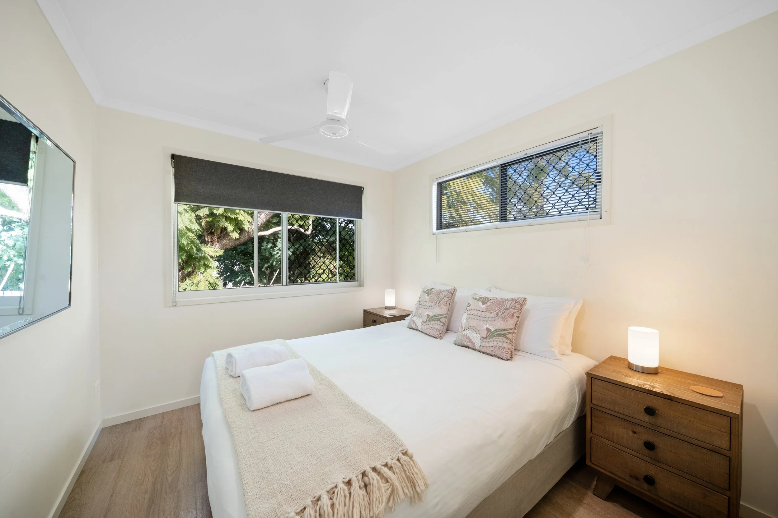 A bedroom with a white bed, two pillows, wooden nightstands, a white lamp, and windows with black blinds and a view of trees outside.