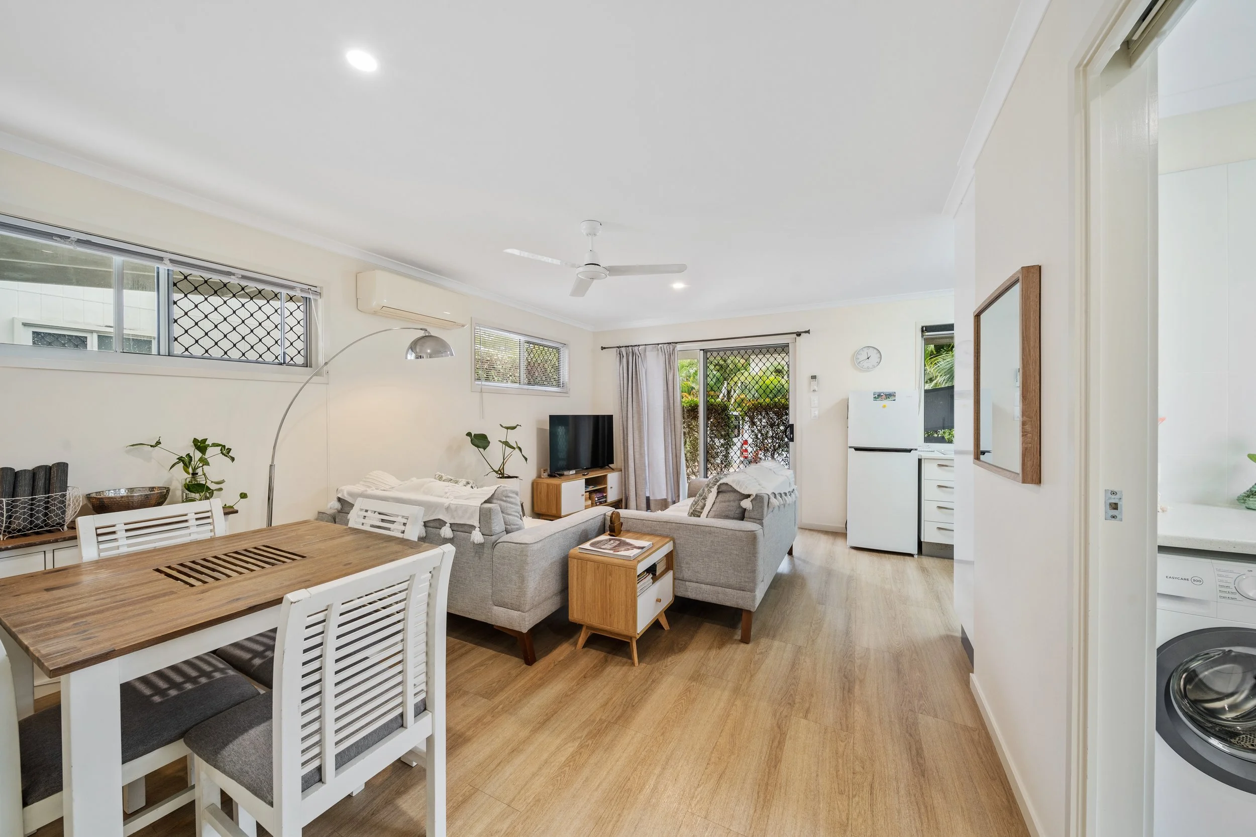 Living room with beige sofas, a wooden coffee table, a television, large windows with curtains, and a dining table with white chairs in an open floor plan.