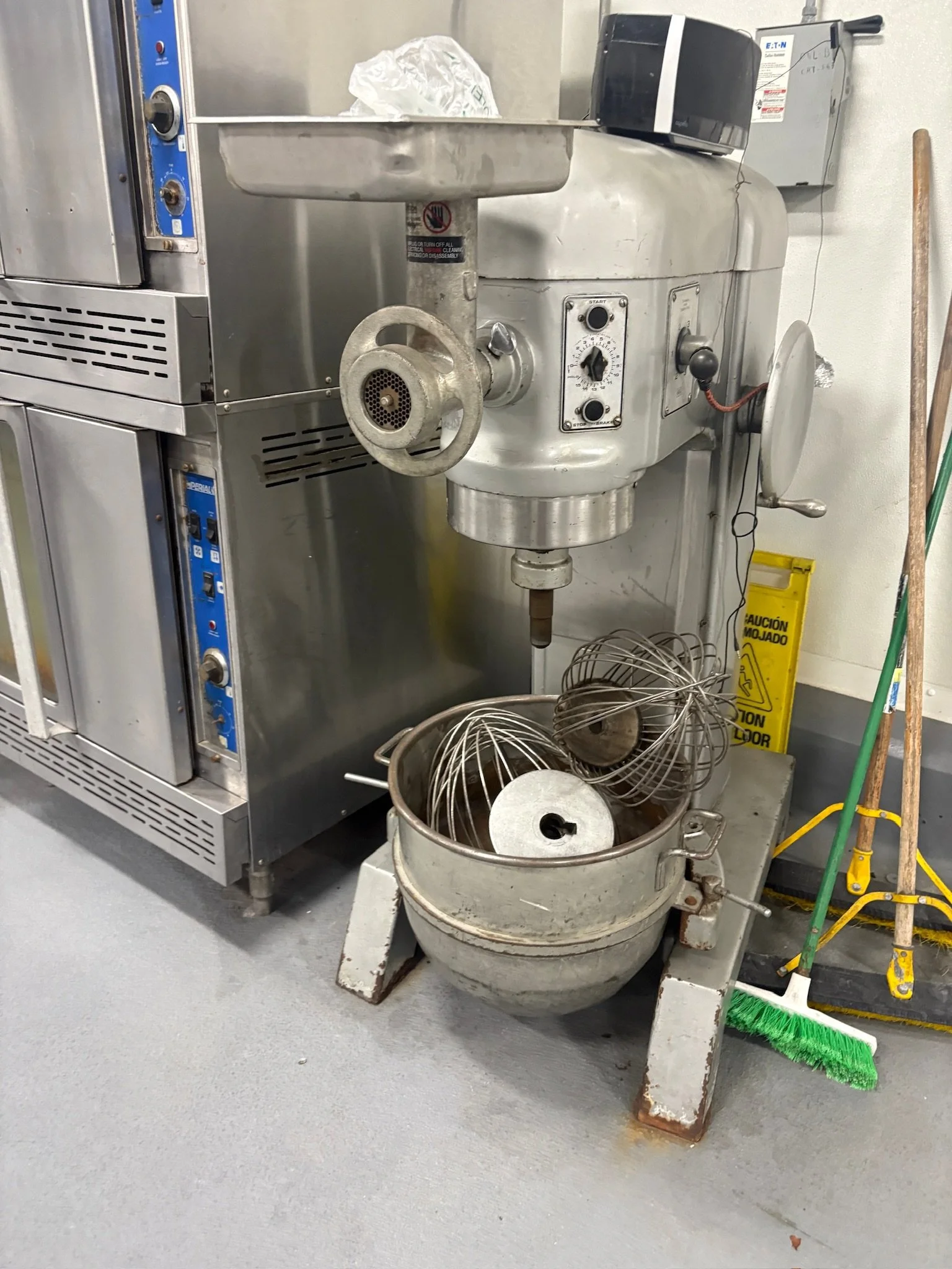 Industrial kitchen equipment including a large grey meat grinder with a circular tray on top, a wire basket underneath, and control knobs on the side. Nearby, there are cleaning tools including a yellow caution sign, a broom with a green brush, and a mop with a green handle. The equipment is situated against a grey wall and floor.