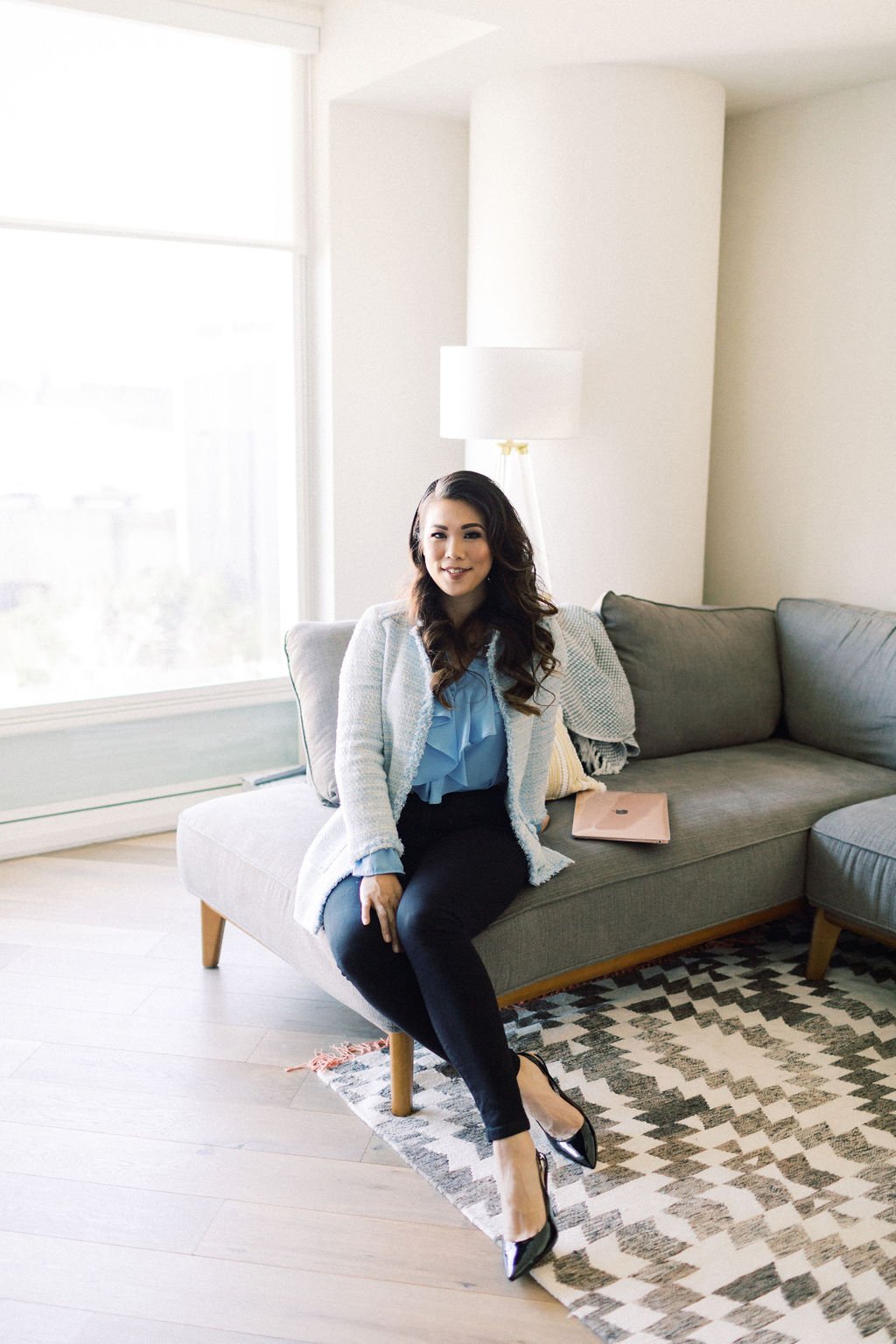 Crystle sitting on a gray couch in a bright living room with large windows, wearing a light blue blouse, black pants, and black high heels, with a pink laptop on the couch beside her.