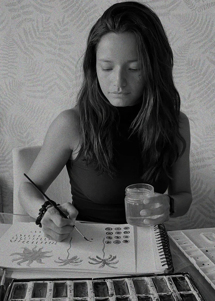 A woman painting a set of three stylized spiders on paper, holding a container of water, with a watercolor palette nearby.