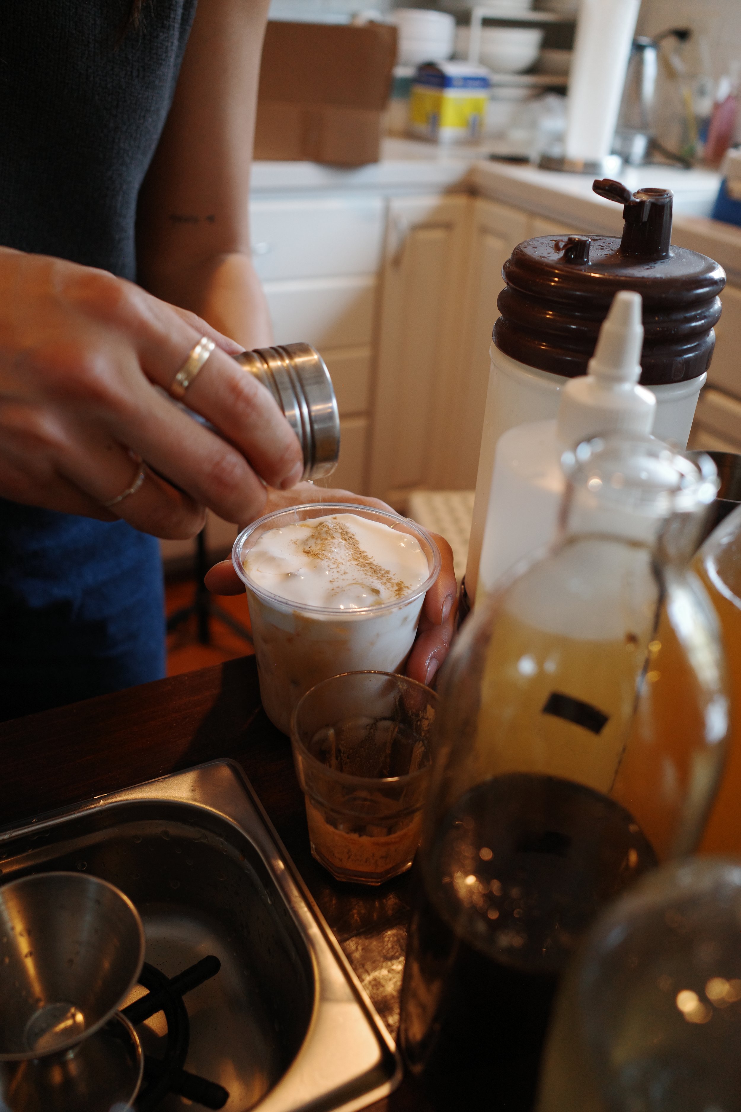 Person pouring caramel syrup into a coffee drink at a kitchen counter with coffee syrups and utensils.