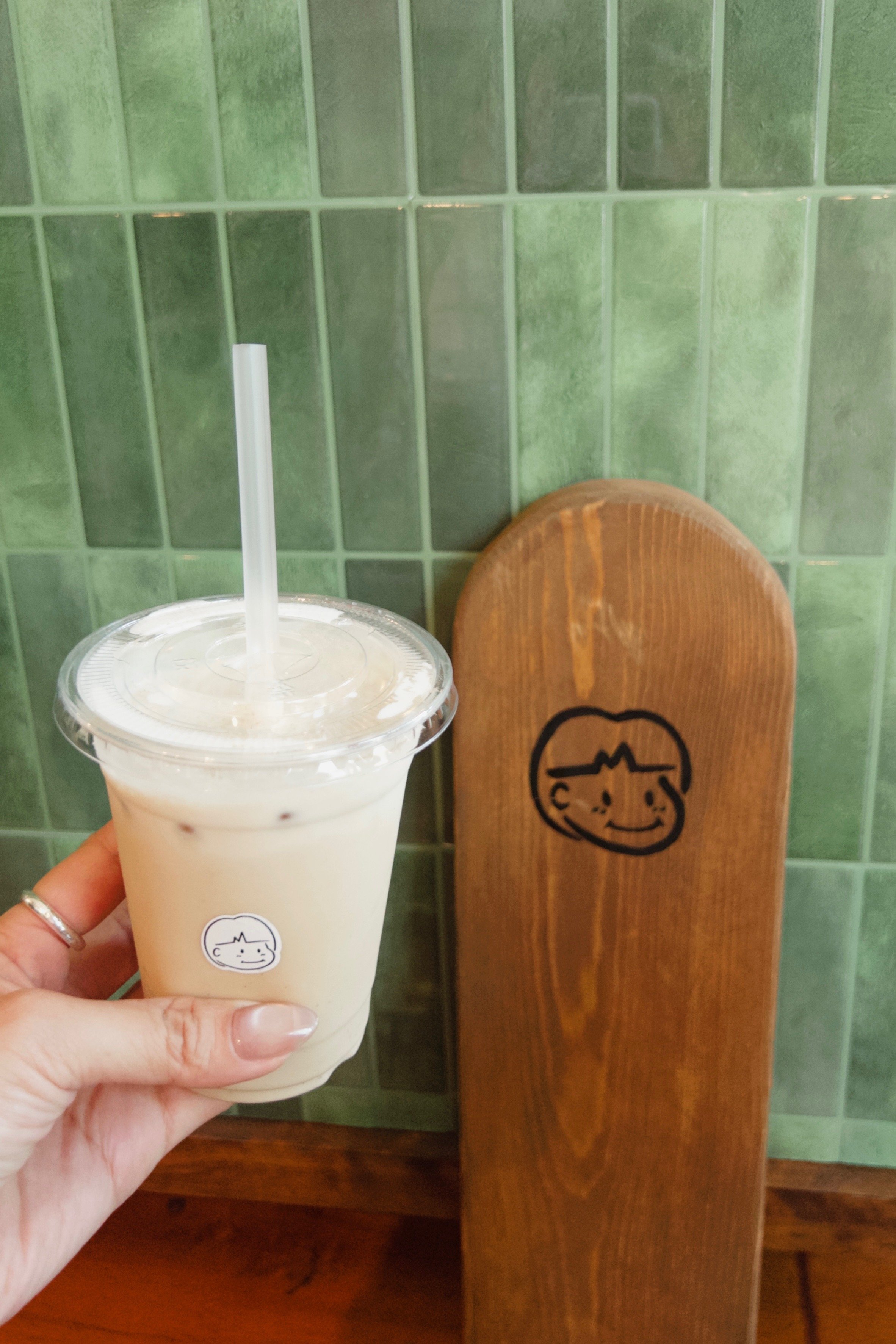 A clear plastic cup with a white beverage and a straw, held by a person with a wedding band. In the background, a wooden chair and green tiled wall with a logo or design. The logo on the cup and chair features a smiling cartoon face with a heartbeat line.