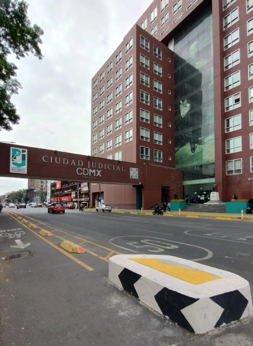 Edificio de la Ciudad Judicial en CDMX con cartel y señales de tráfico, y un muro decorado con un mural grande de un rostro.