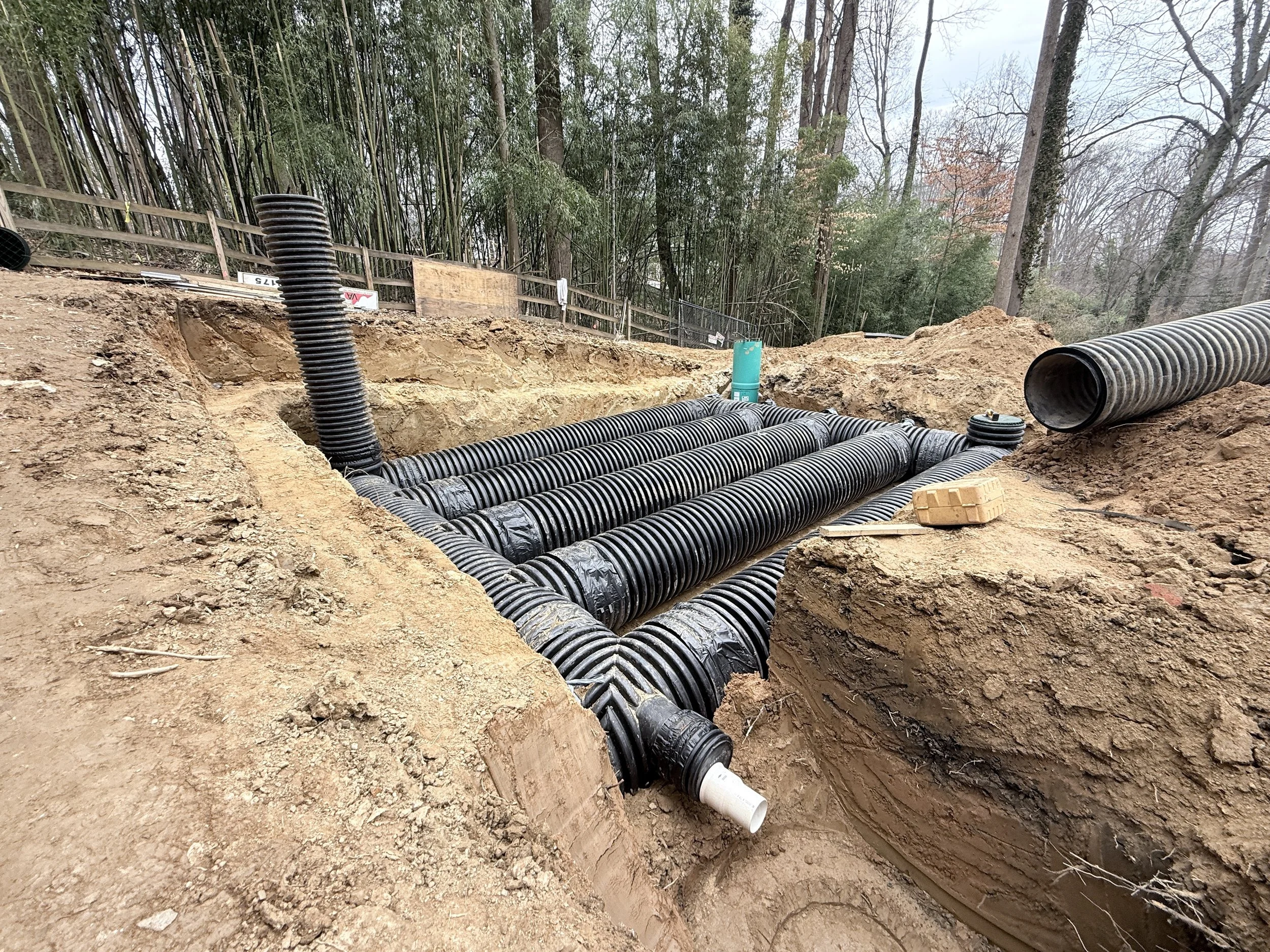 Underground construction site with black corrugated pipes installed in trenches, surrounded by a wooded area with trees and bushes.