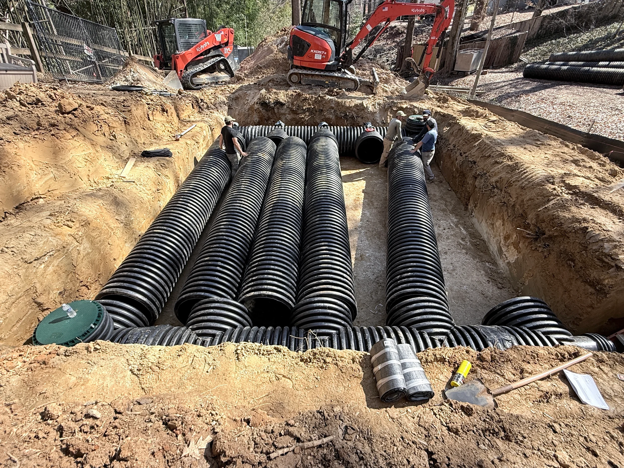 Crew installing HDPE underground detention pipes in a large excavation with corrugated pipe runs, access risers, and site equipment.