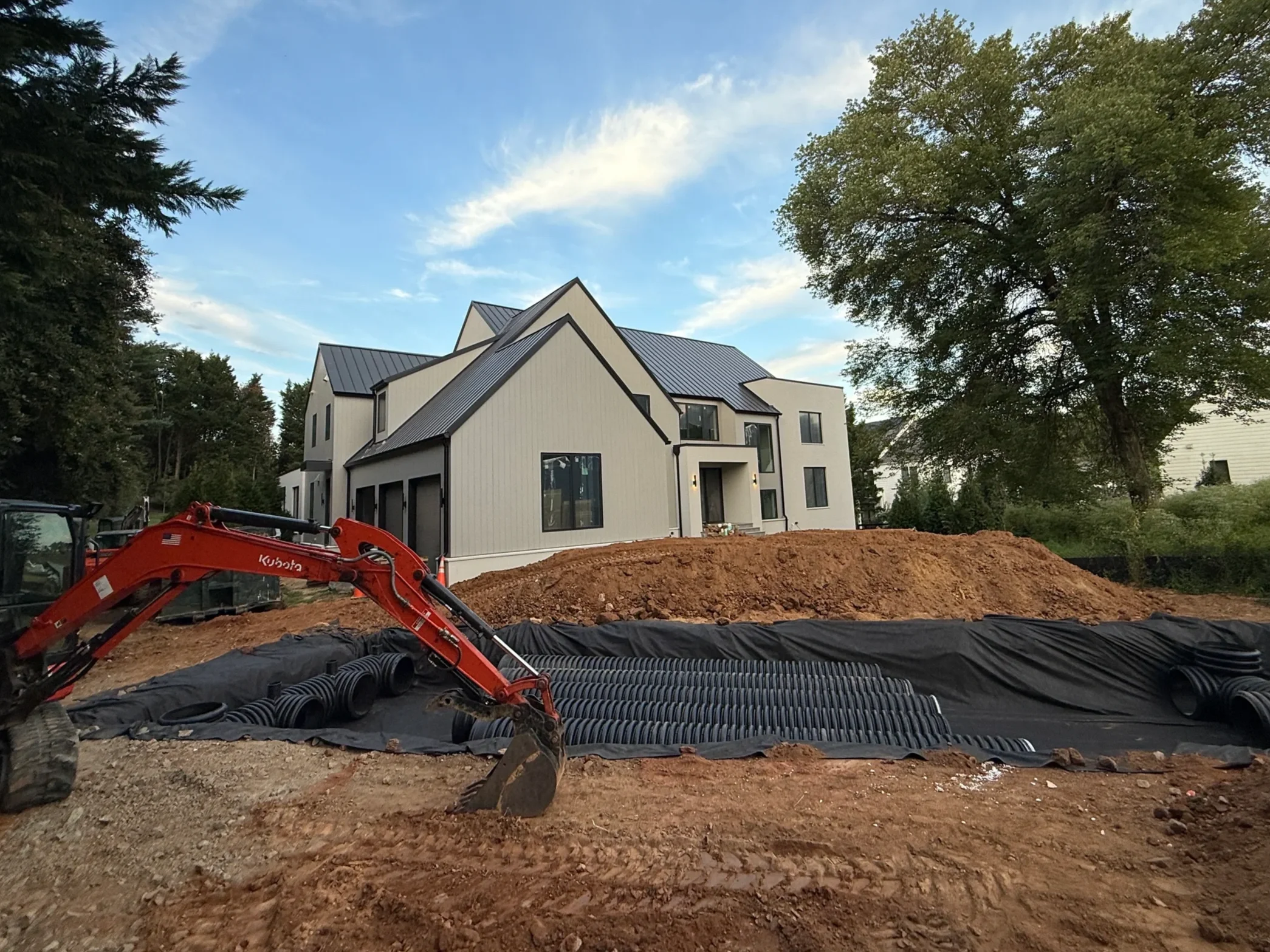Residential underground pipe detention installation beside a home with excavator, corrugated HDPE pipe runs, geotextile liner, and graded soil.