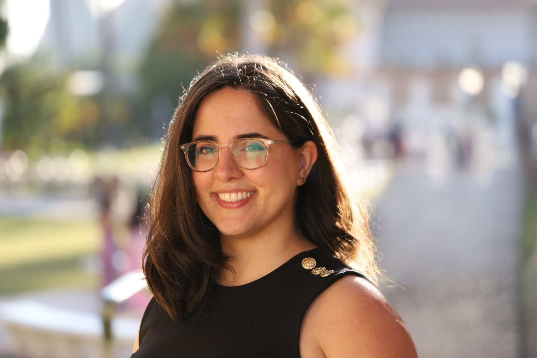 A woman with brown hair, glasses, and a black sleeveless top smiling outdoors on a sunny day, with blurred trees and a playground in the background. She has a warm smile.