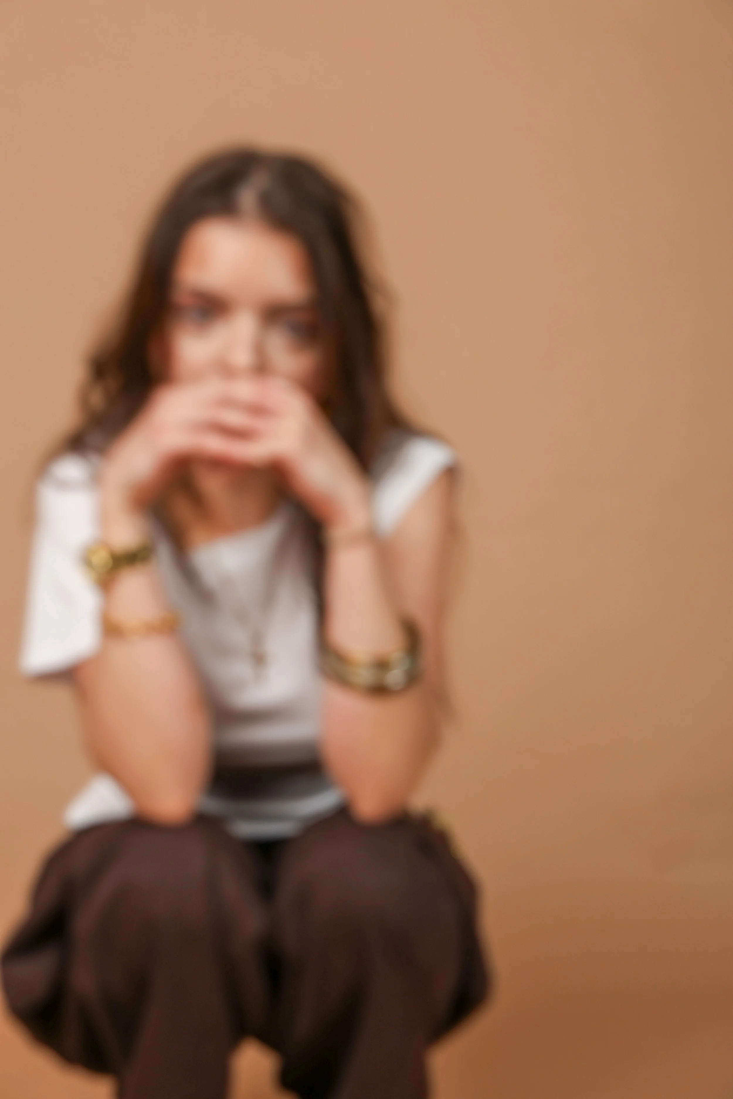 A woman with dark hair sitting with her arms resting on her chin, wearing a white top and jewelry, against a plain beige background.