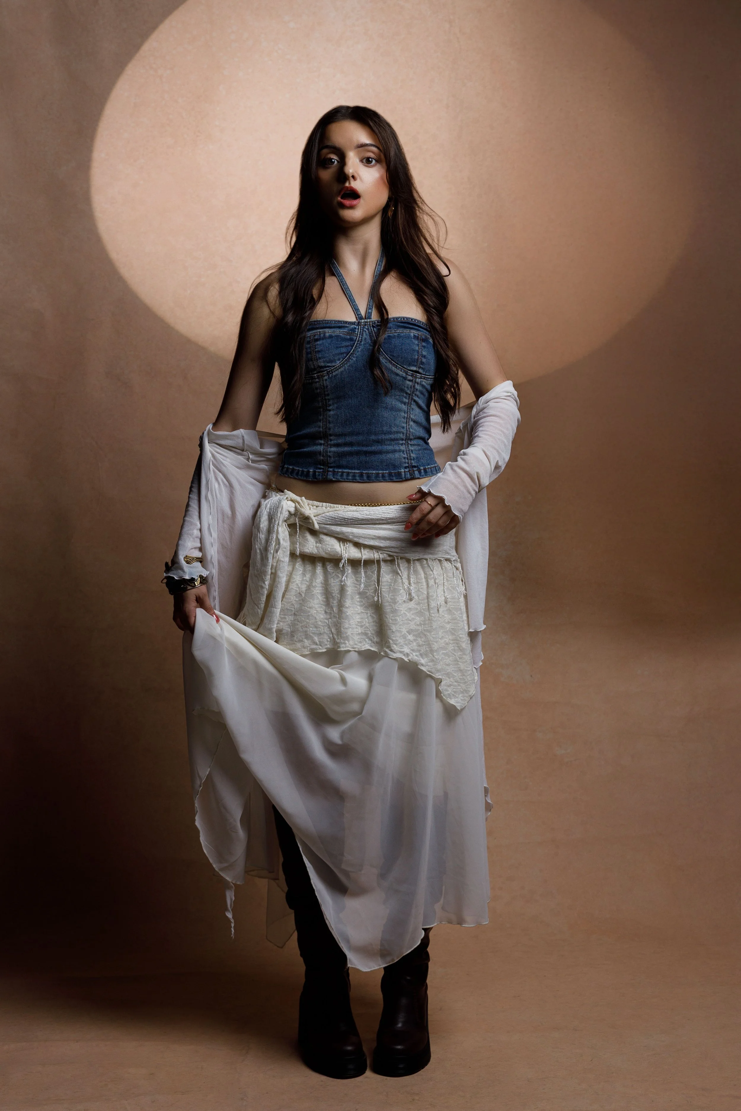 A young woman with long dark hair wearing a denim bustier top, layered white skirt, and black boots, standing in front of a beige background with a large spotlight circle behind her.