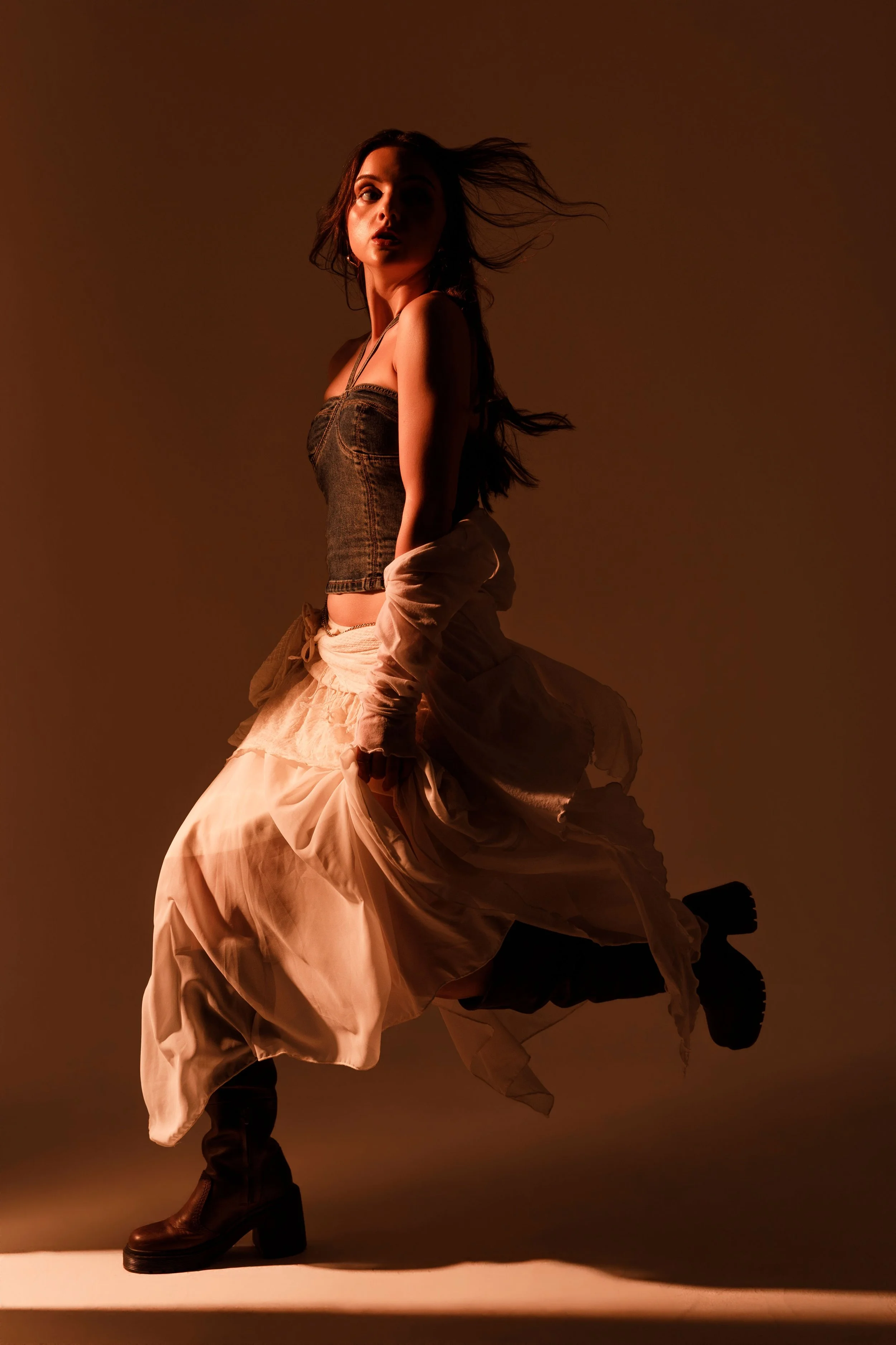 A woman with long hair, wearing a dark sleeveless top, is standing against a dark background. She is dressed in flowing beige skirt or coat and black boots, with her hair blowing. The lighting highlights her face and outfit.