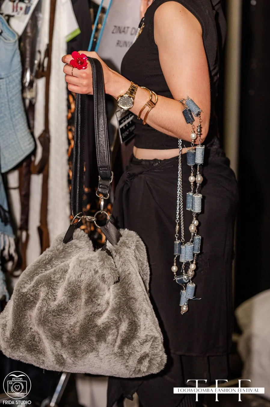 Close-up of a woman's arm holding a plush gray bag with a black strap at Toowoomba Fashion Festival, showcasing jewelry including a watch, bracelets, and a long necklace with denim-like beads and pearls.