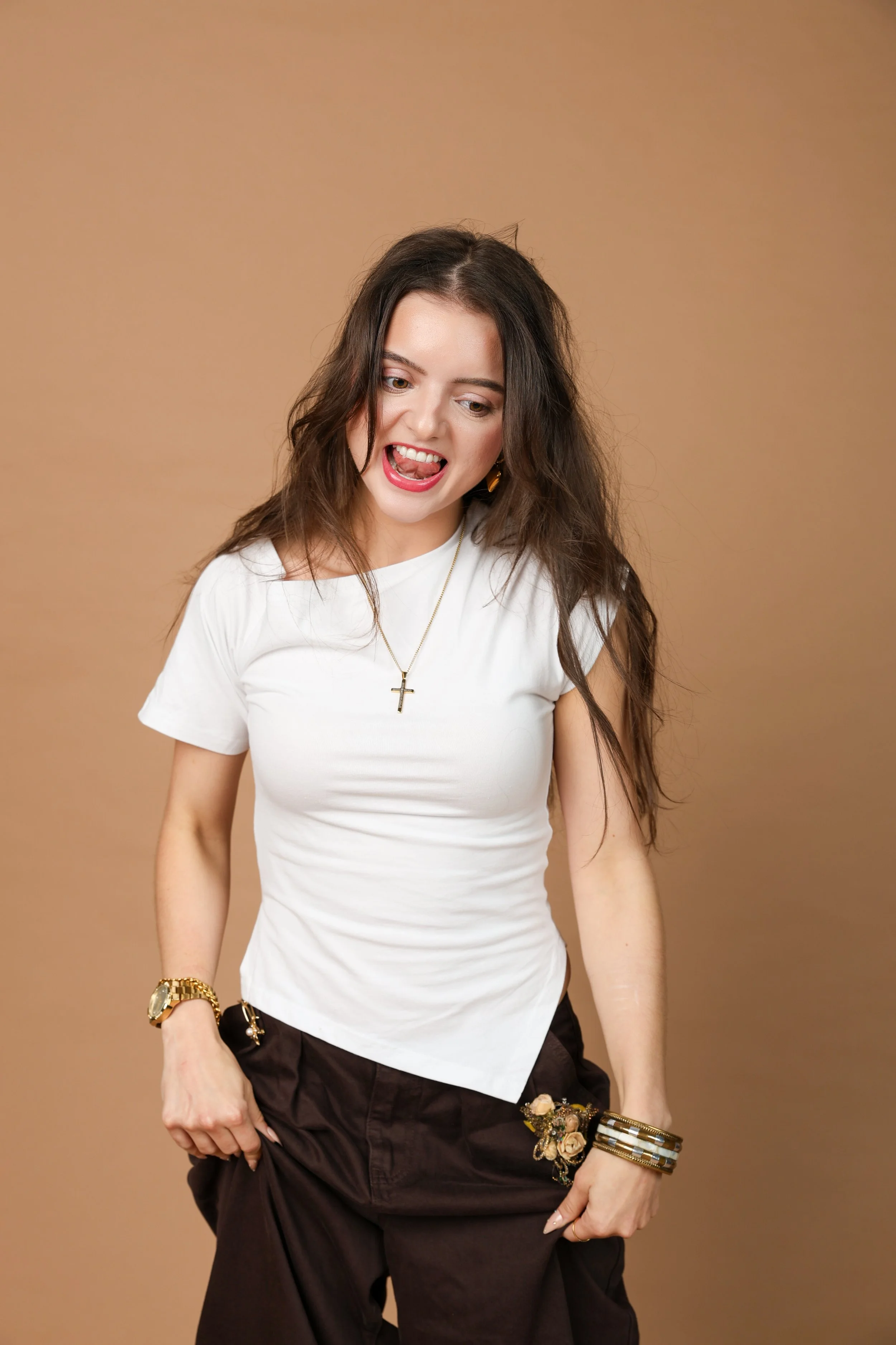 A woman with long wavy brown hair looks down and sticks out her tongue, wearing a white t-shirt with a cross pendant necklace, gold watch, black shorts, and layered bangles, standing against a plain beige background.