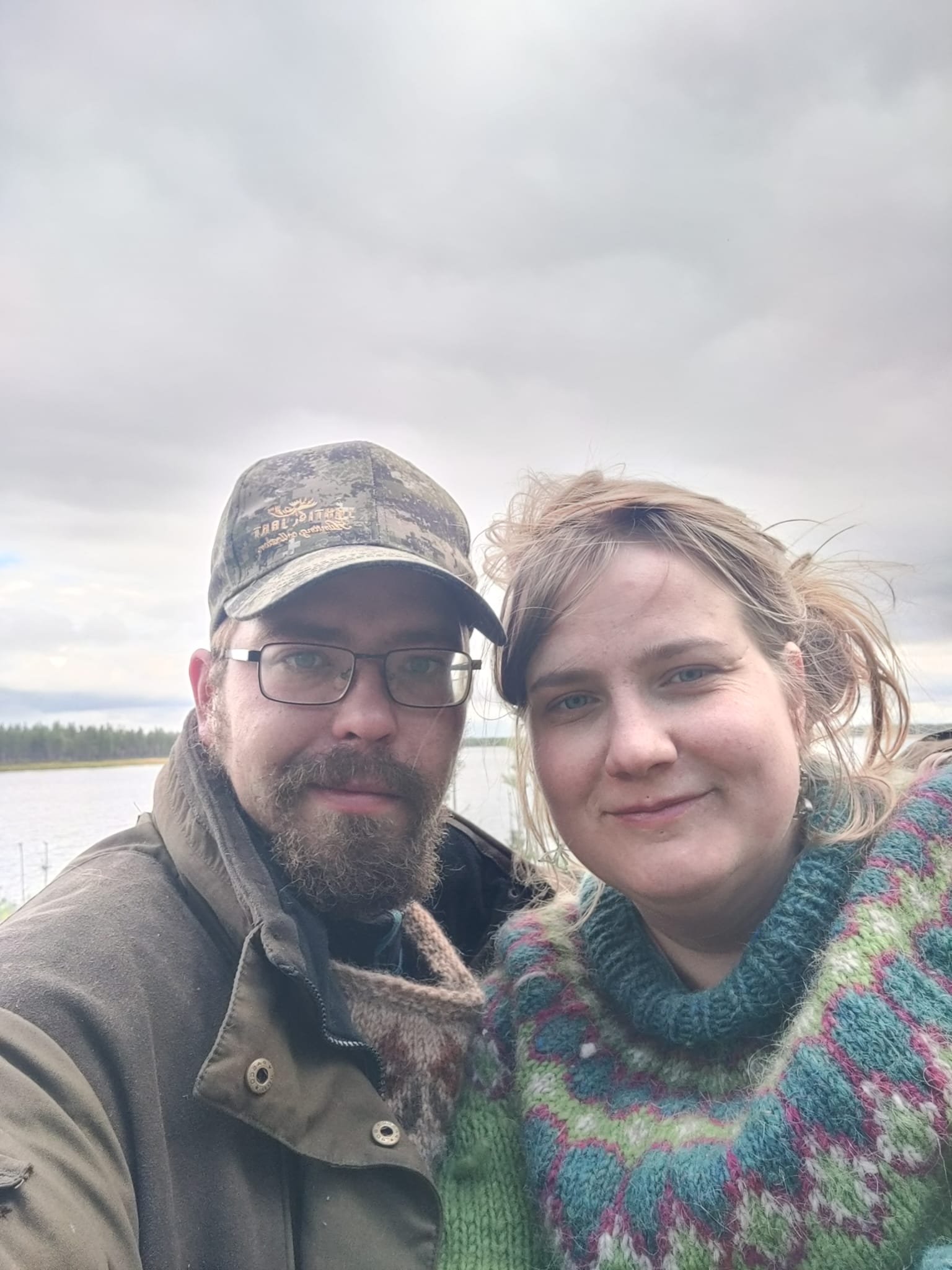 A close-up photo of a man and a woman outdoors with a cloudy sky and body of water in the background.