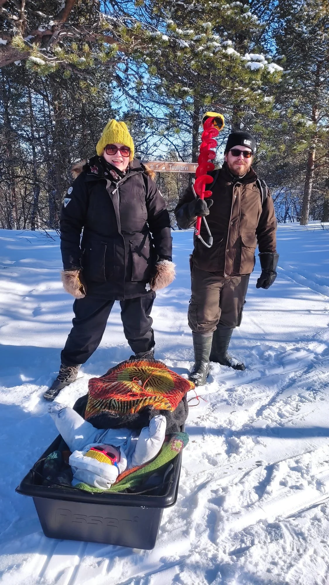 Two people stand in a snowy forest holding an ice rescue tool, with a child lying in a sled in front of them. They are dressed in winter clothing, including gloves, hats, and sunglasses.
