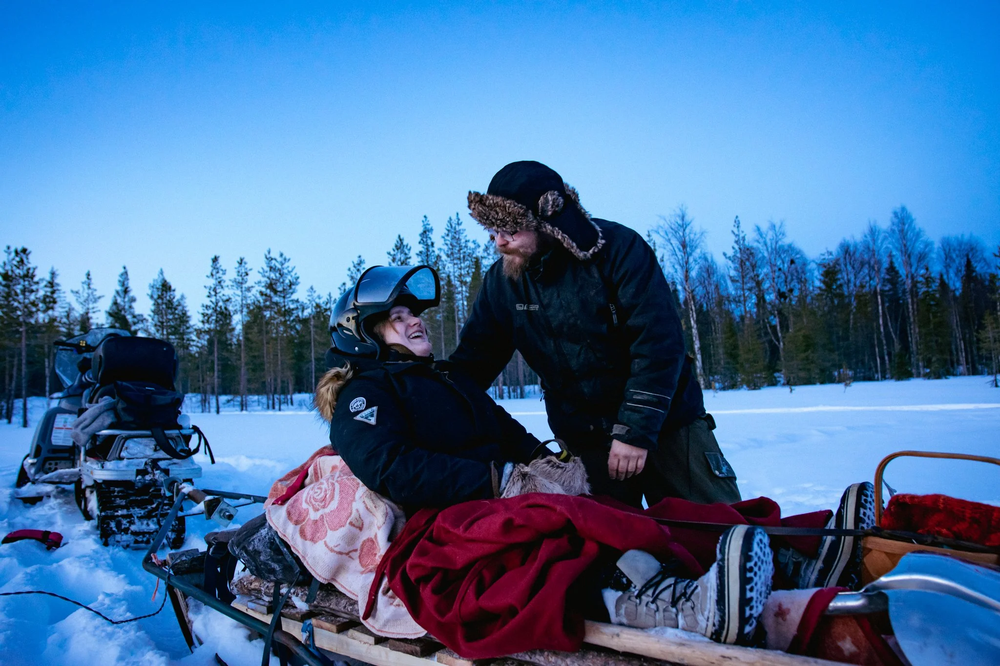 A woman sitting on a sled, smiling at a man who is standing and leaning towards her, in a snowy outdoor setting with a forest of trees in the background. There is a snowmobile and winter gear around them.