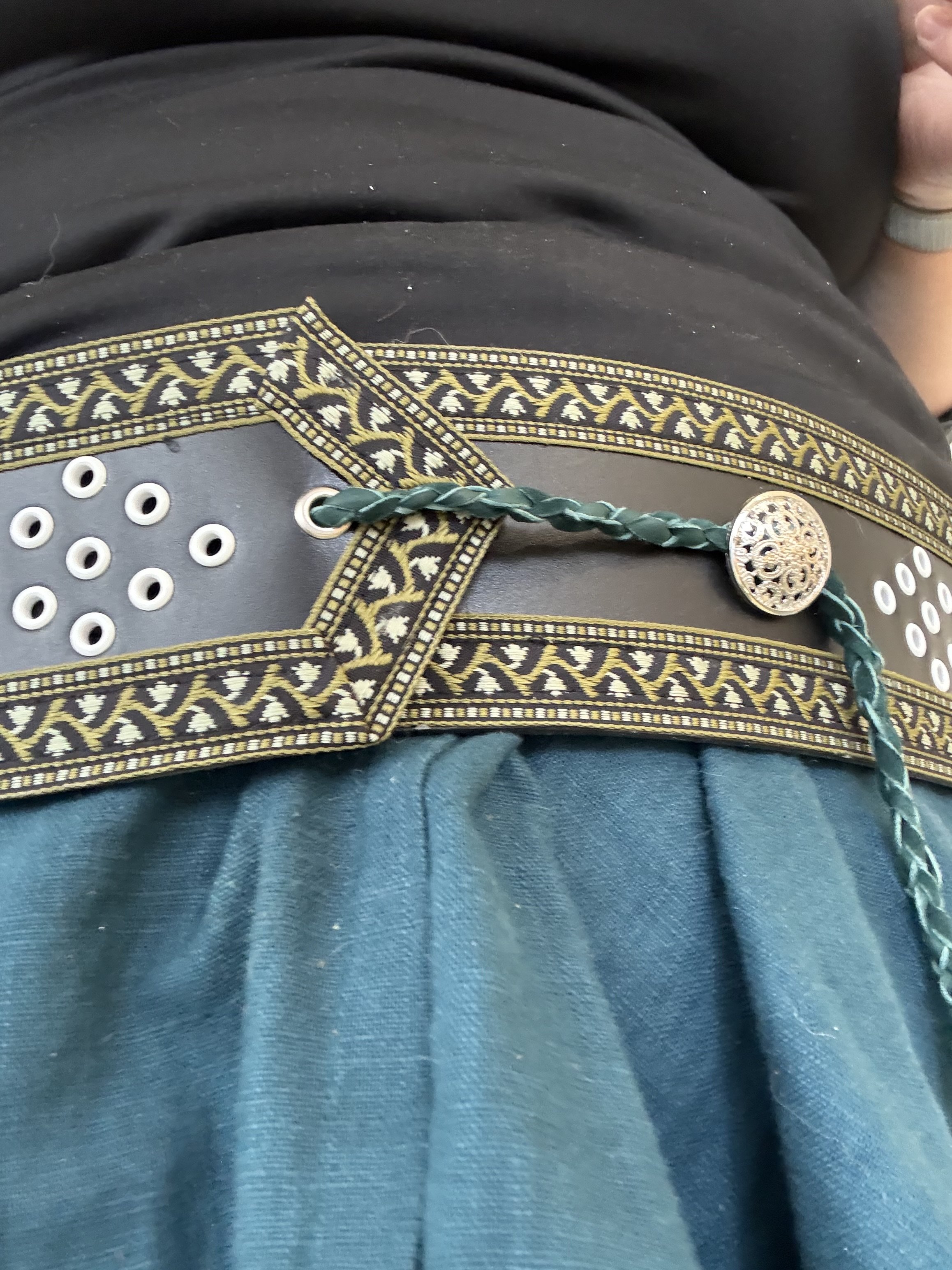 Close-up of a person's belt and lower shirt with decorative trim, eyelets, and a braided green belt with a round metallic buckle, worn over blue pants.
