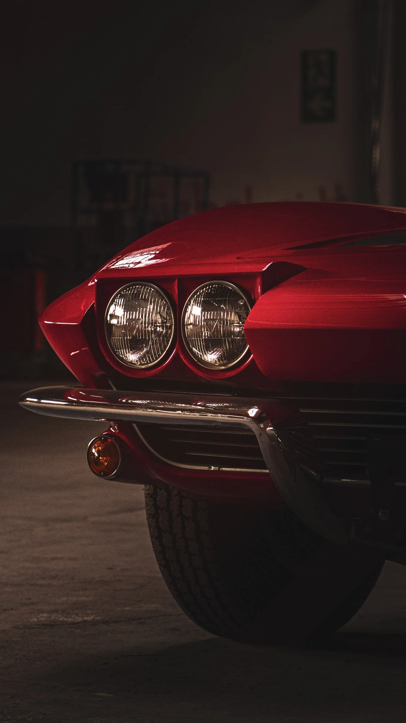 Teil der Front einer roten Corvette, mit runden Scheinwerfern, Scheinwerferabdeckungen aufgeklappt und Chromstoßstange, in einer dunklen Halle.