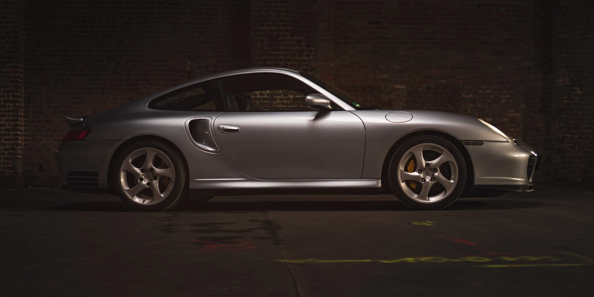 Silberner Porsche 911 in einer dunklen Garage vor einer Ziegelwand.