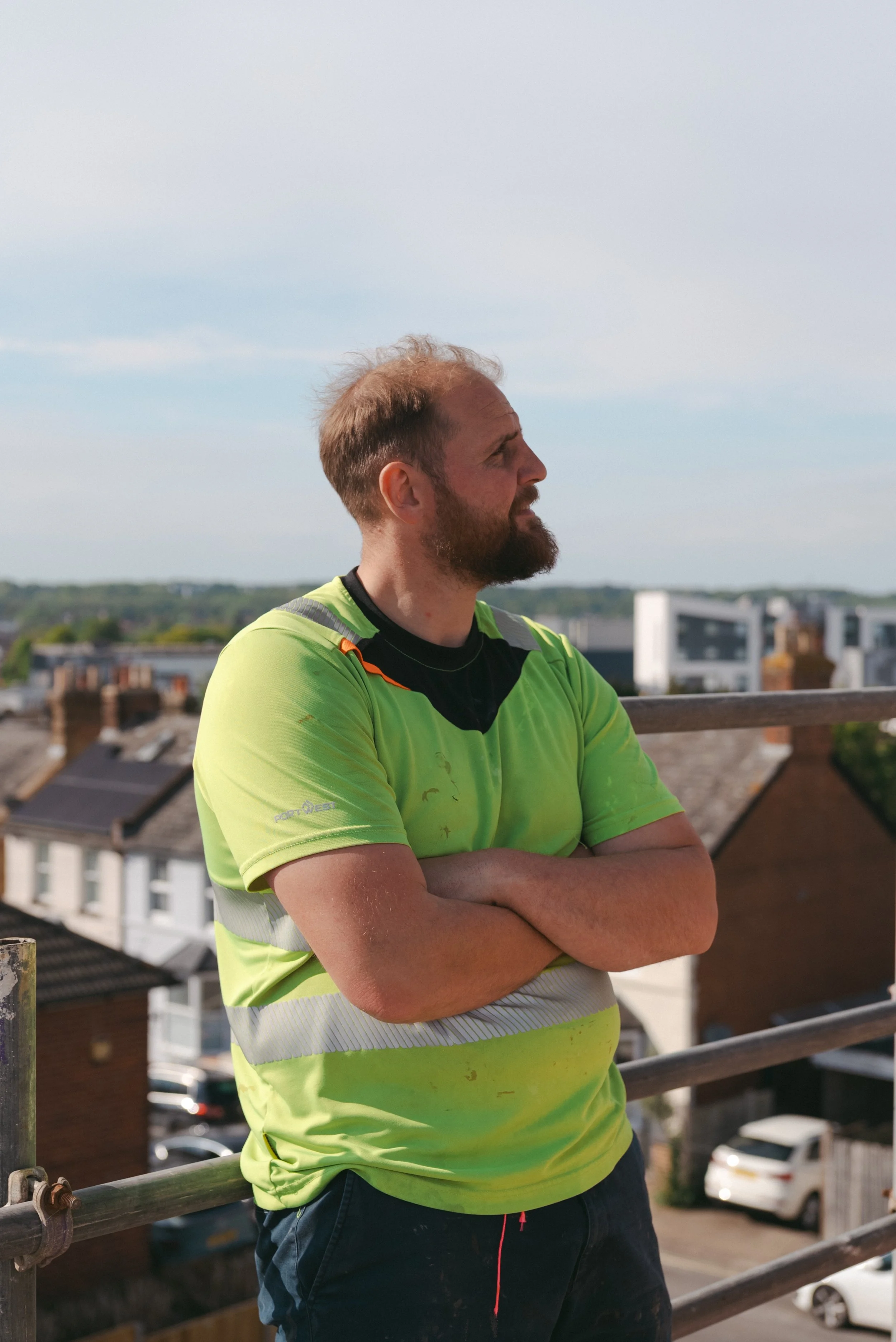 A man with a beard and short hair, wearing a bright yellow safety shirt, stands outdoors on a rooftop with houses and cars in the background, crossing his arms and looking to the side. Builders, building, carpenters, carpentry, East Kent, trust
