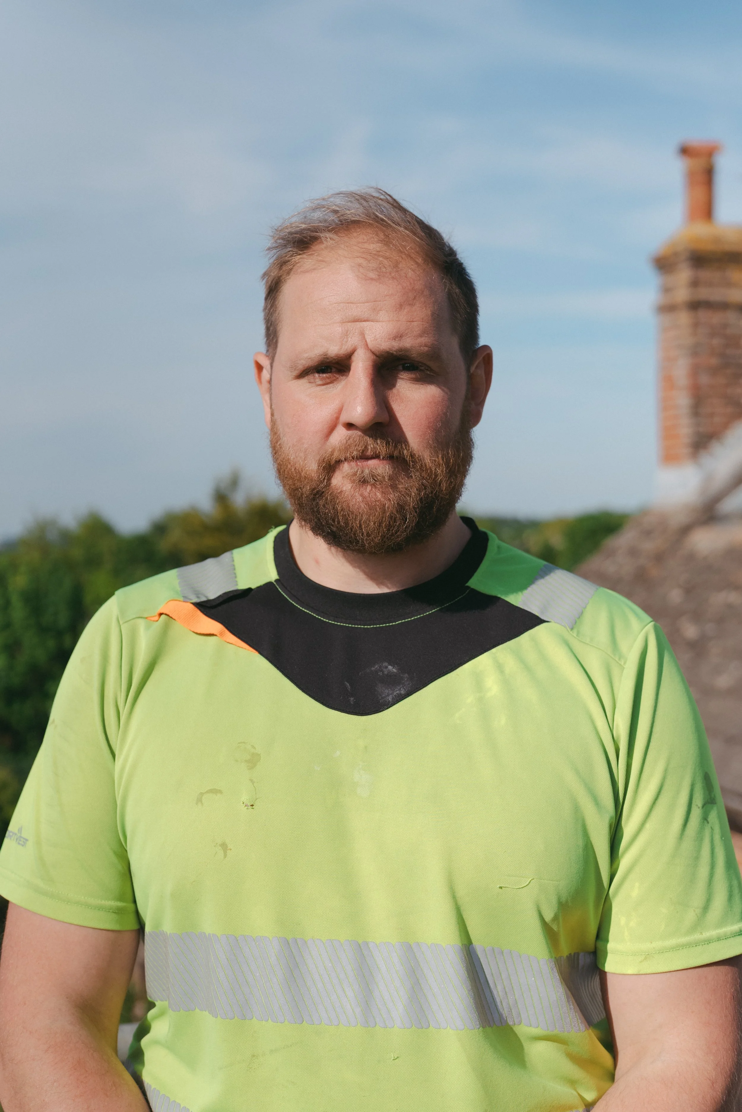 A man with a beard and short hair wearing a bright yellow green athletic shirt with black and orange accents, standing outdoors under a blue sky with a brick chimney in the background. Builders, building, carpenters, carpentry, East Kent, trust