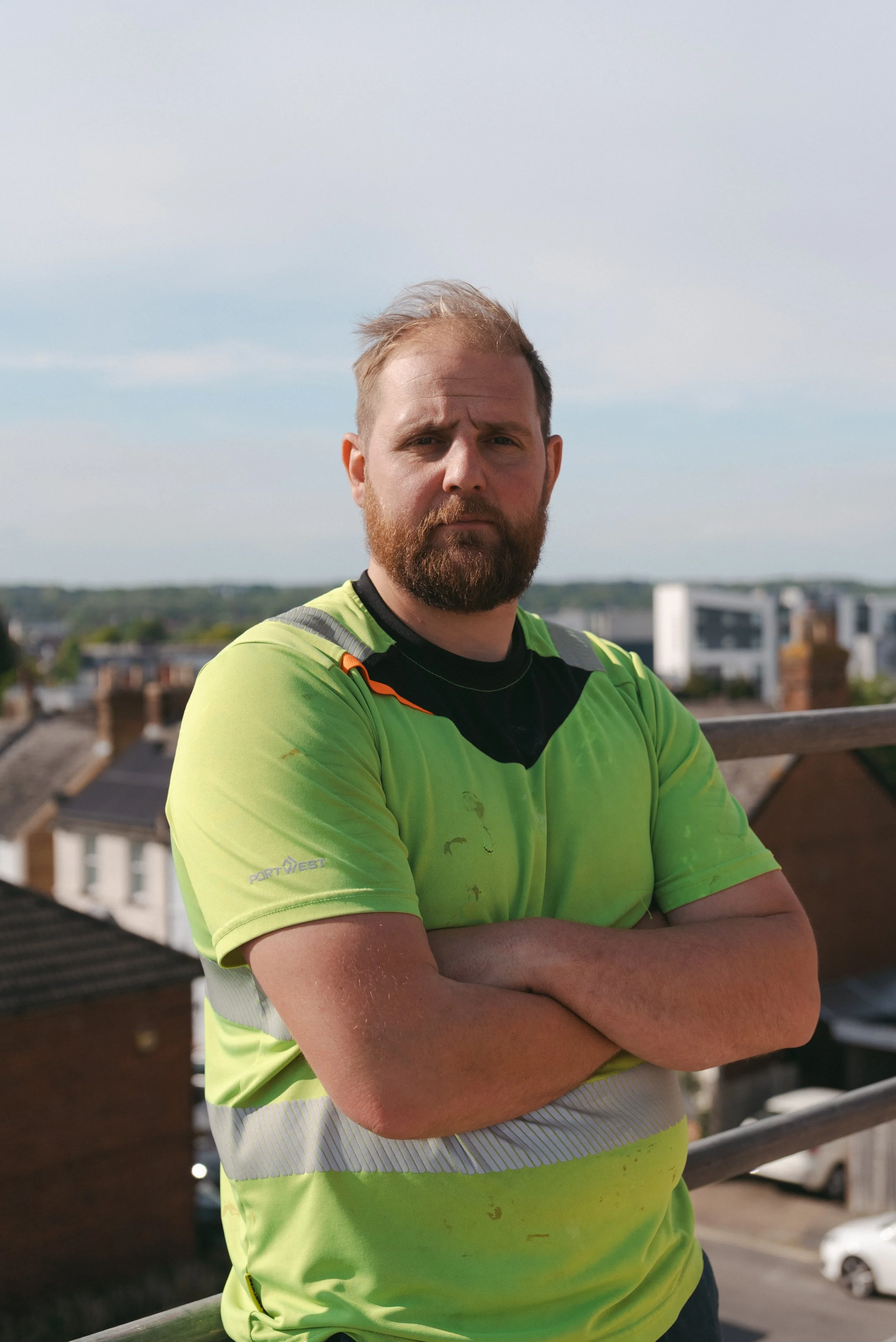 A man with a beard and light brown hair standing outdoors on a rooftop, wearing a bright yellow and black sports shirt, with houses and buildings in the background under a partly cloudy sky. Builders, building, carpenters, carpentry, East Kent, trust