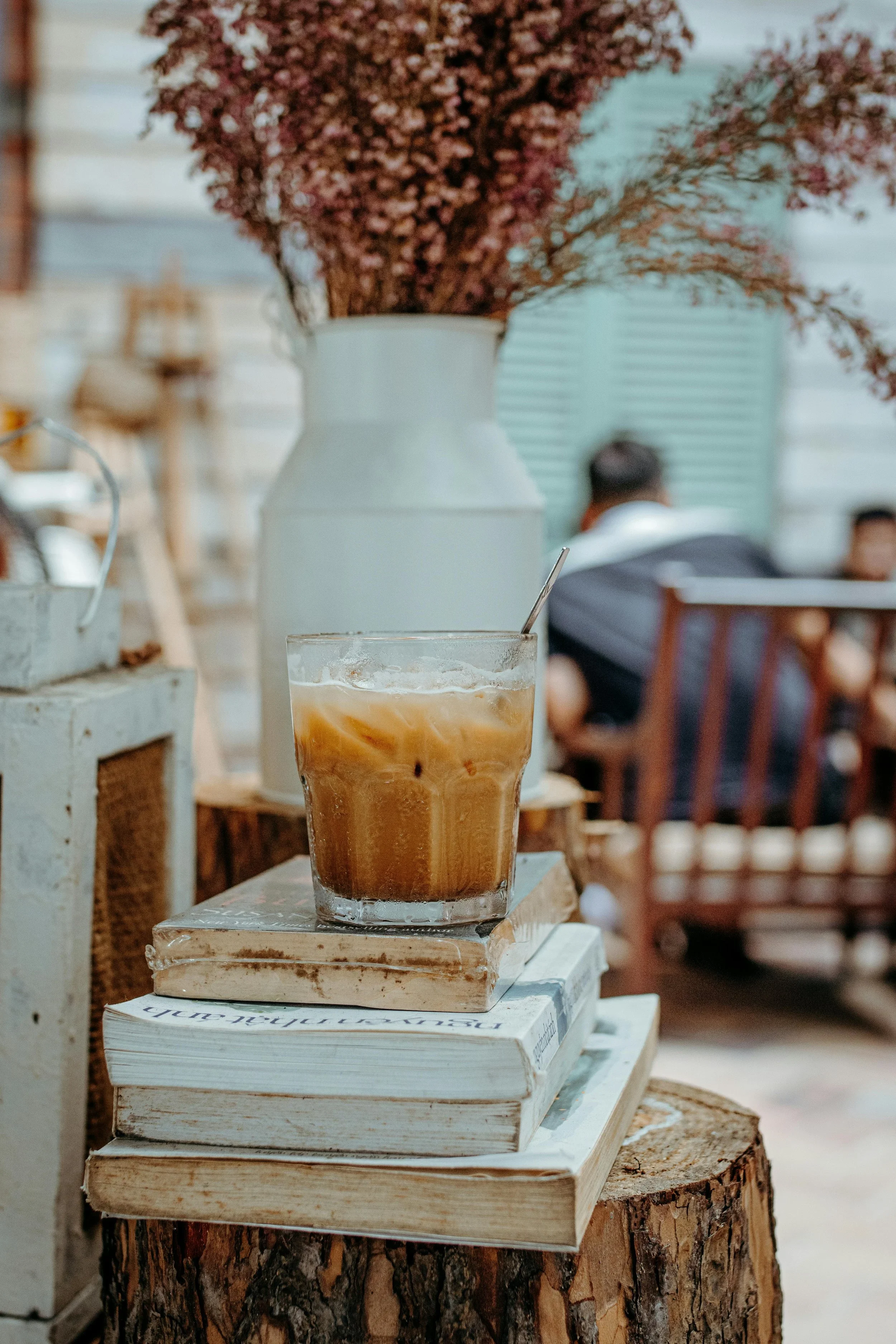 Een glas iced coffee op een stapel boeken, geplaatst op een stuk boomstam met een witte vaas met gedroogde bloemen op de achtergrond.