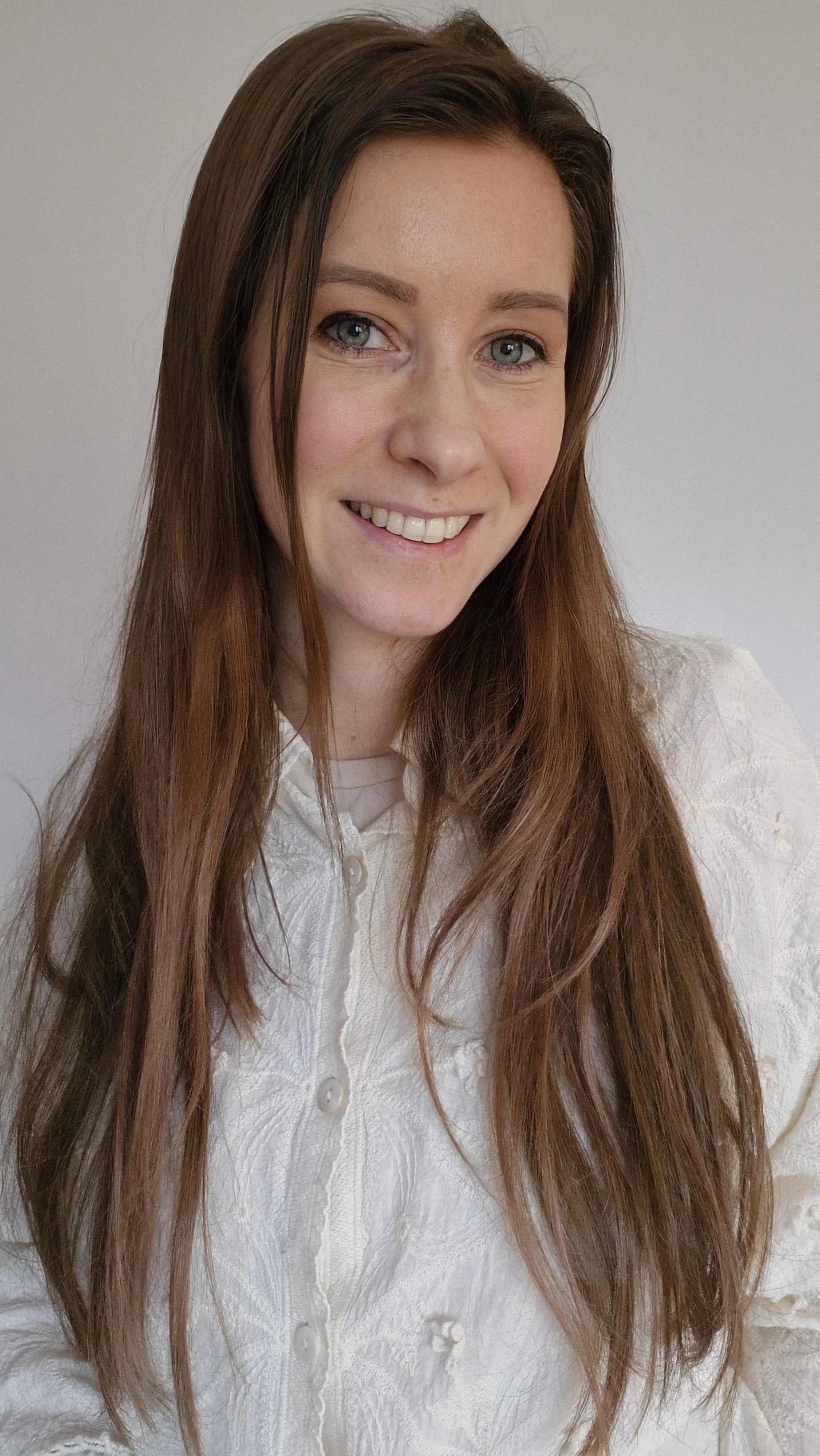 Portret van een vrouw met lang, kastanjebruin haar en blauwe ogen, lachend tegen een witte achtergrond.