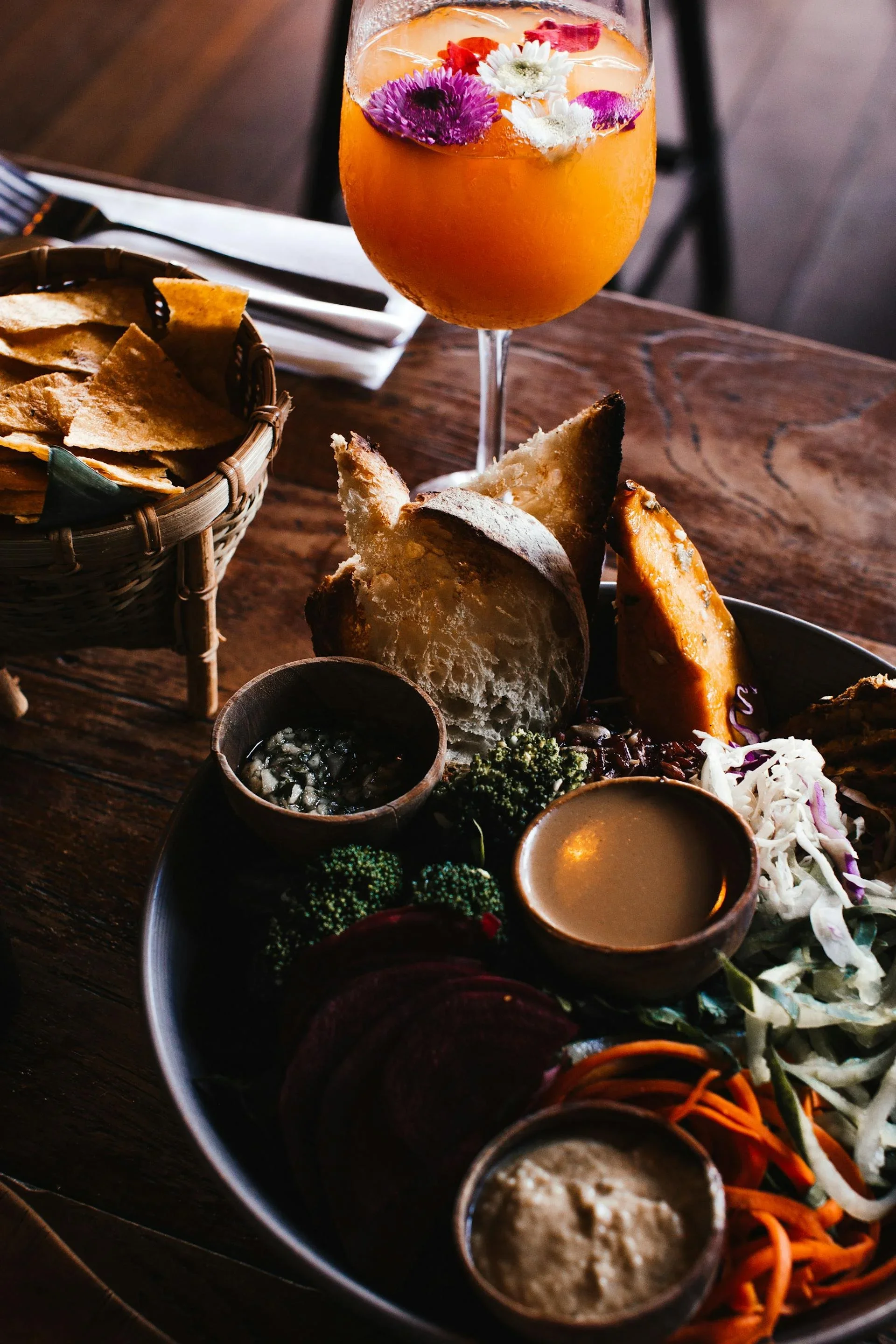 Een plateau met diverse soorten groenten, dips en brood, met een glas met een kleurrijke drank met bloemen en fruit.