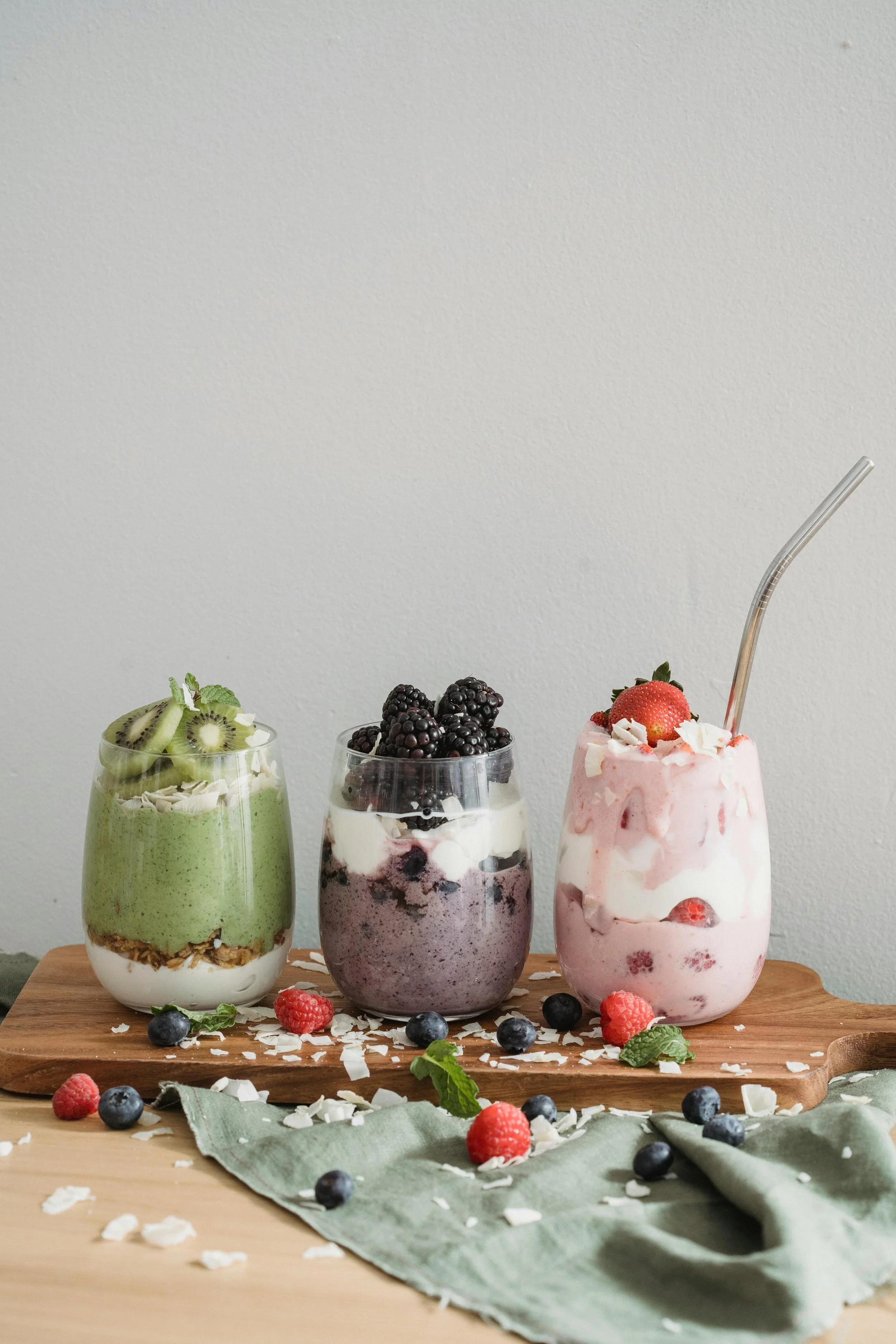 Drie glazen met kleurrijke fruit-yoghurts op een houten snijblok, omringd door vers fruit, meringen en kokosvlokken, op een groene doek.